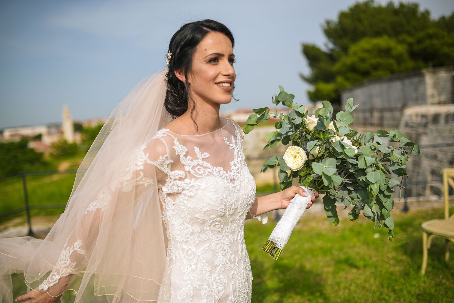 Beautiful bride in lace wedding gown holding elegant white and green bouquet outdoors on sunny day.