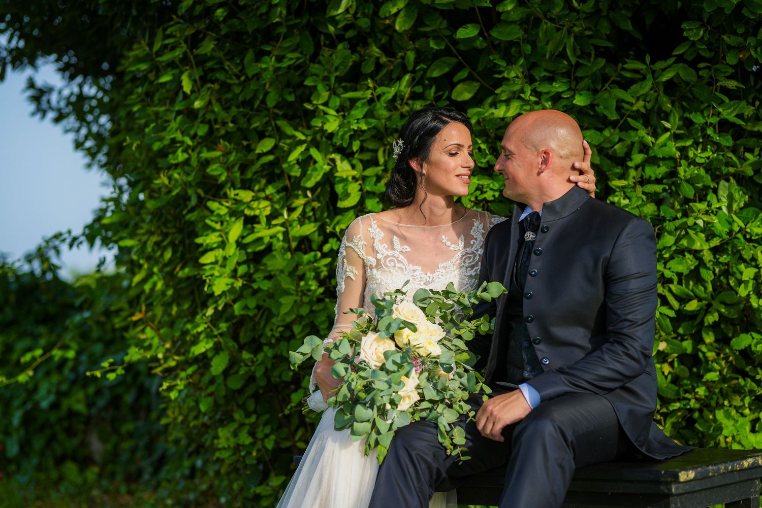 Bride and groom sitting together in front of lush green hedge holding a white floral bouquet.