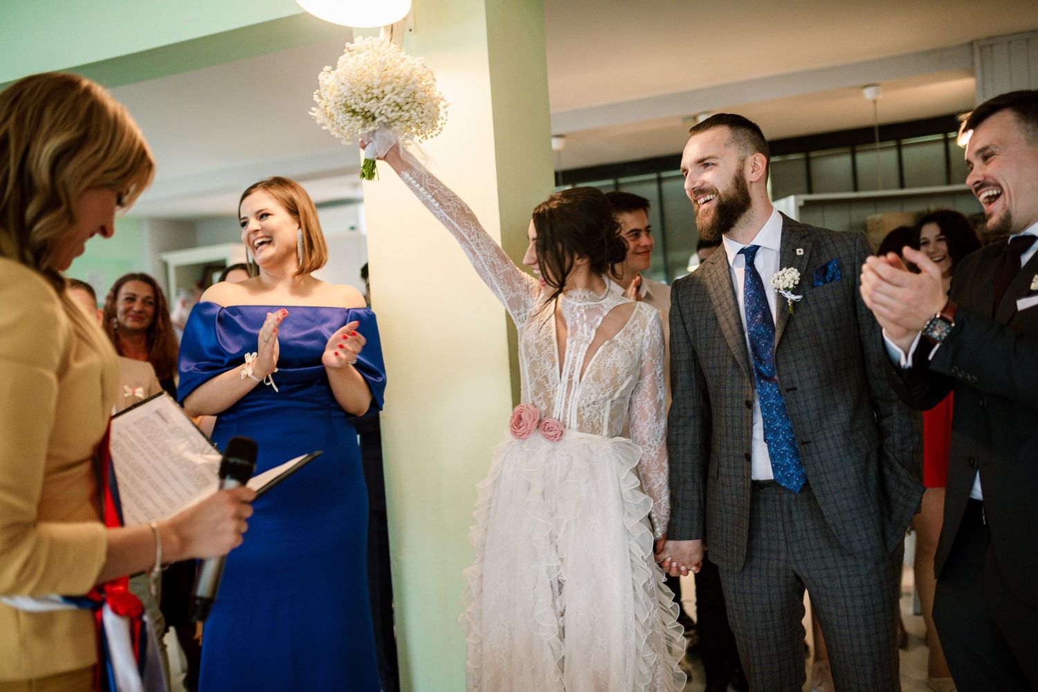 Bride raises bouquet triumphantly while bearded groom smiles during joyful wedding reception.