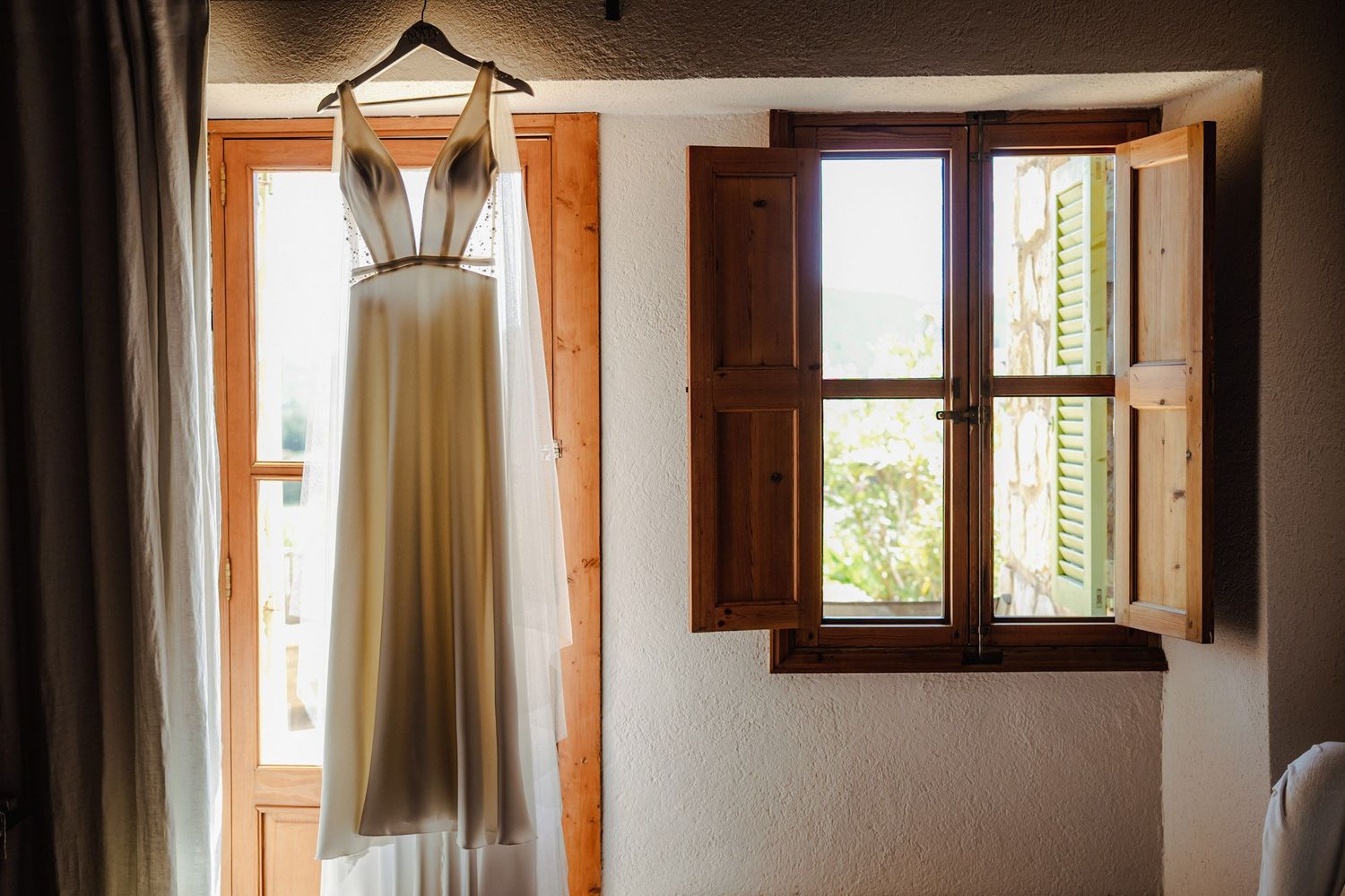 Elegant sleeveless wedding dress hanging on wooden window frame in rustic stone villa room.