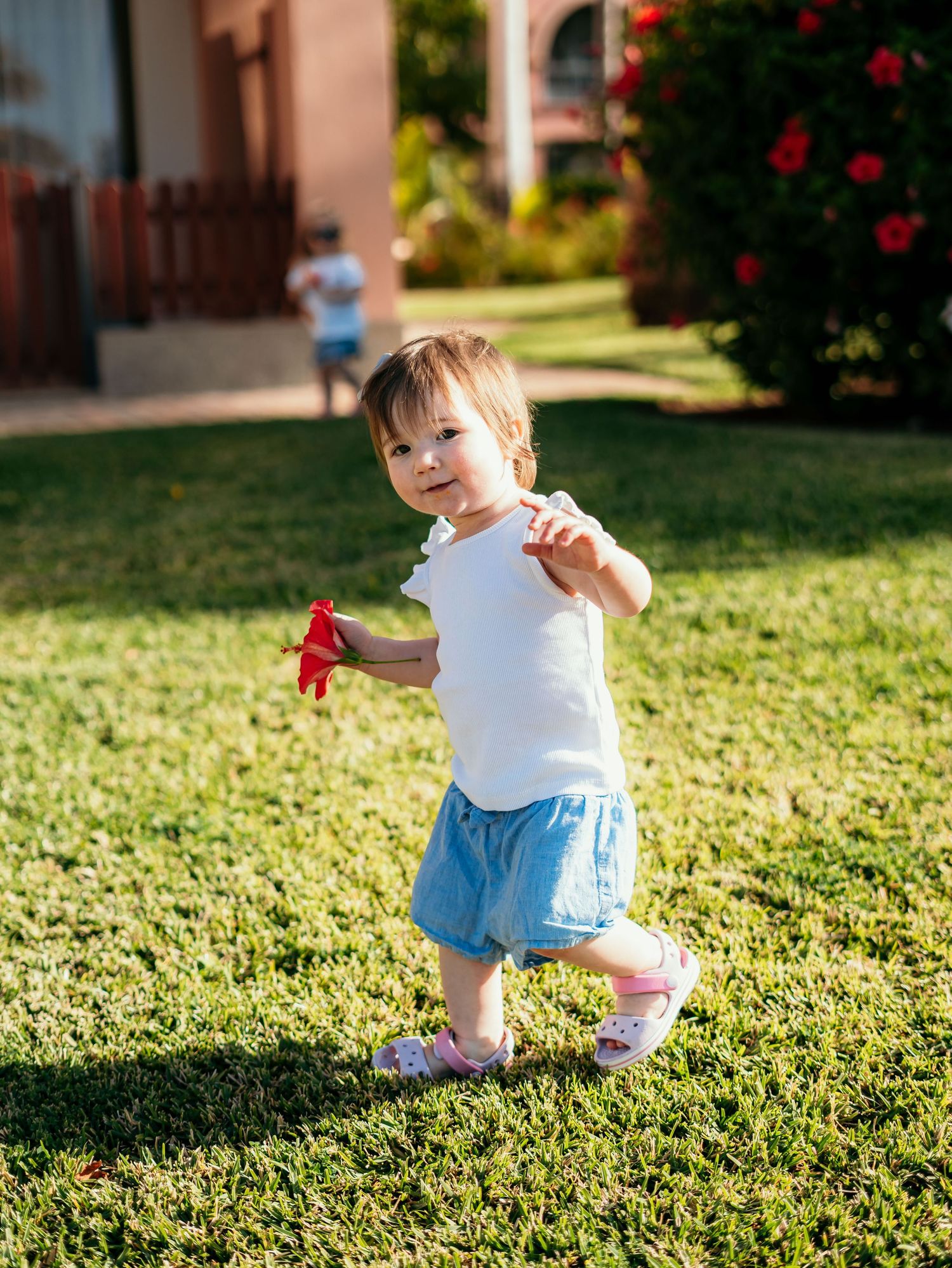 Toddler girl in white top and blue skirt walking on green grass holding a red toy in sunny garden.