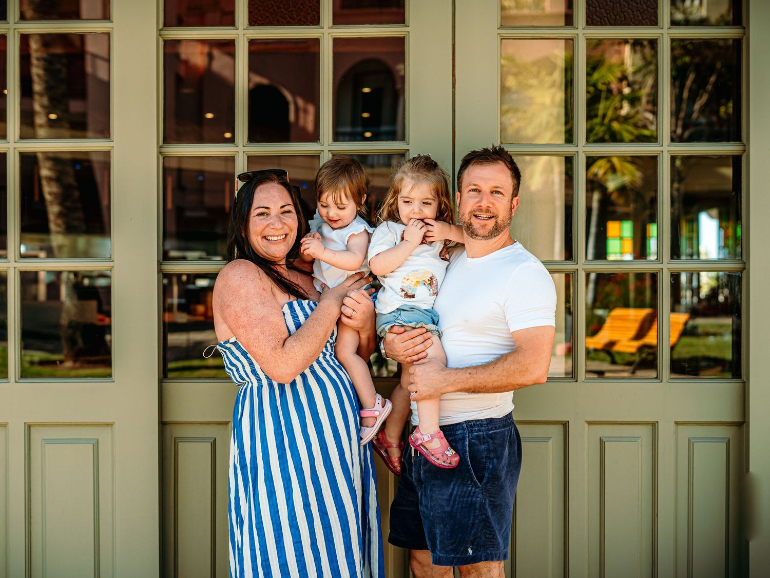Happy family of four with two young daughters posing in front of sage green French doors outdoors.