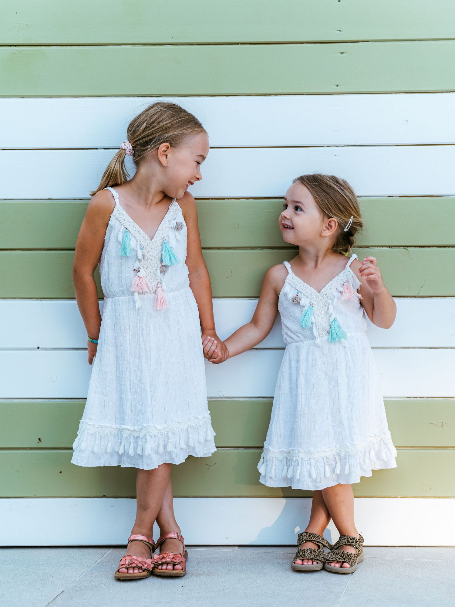 Two young sisters in matching white tassel dresses hold hands and smile at each other by a colorful wall.
