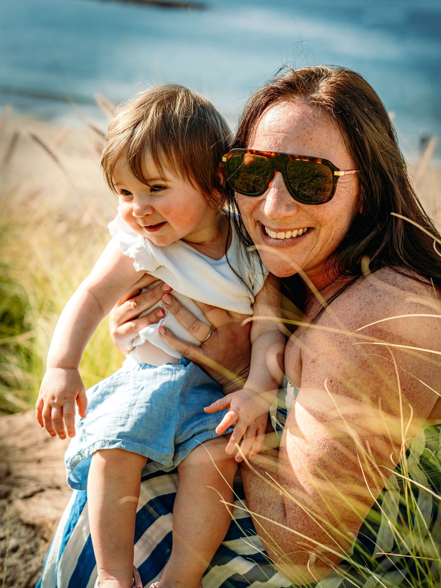 A smiling mother with sunglasses holds her happy toddler on a sunny beach with golden grass.