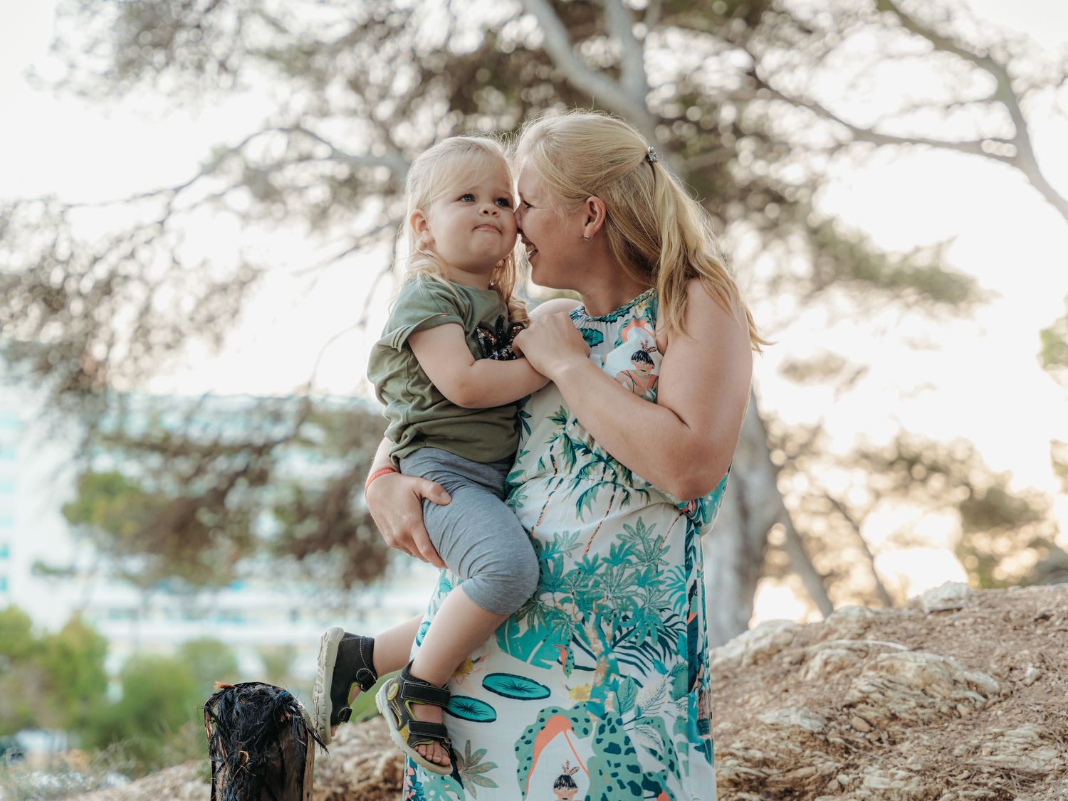 A loving mother in floral dress holds her toddler son close outdoors near pine trees.