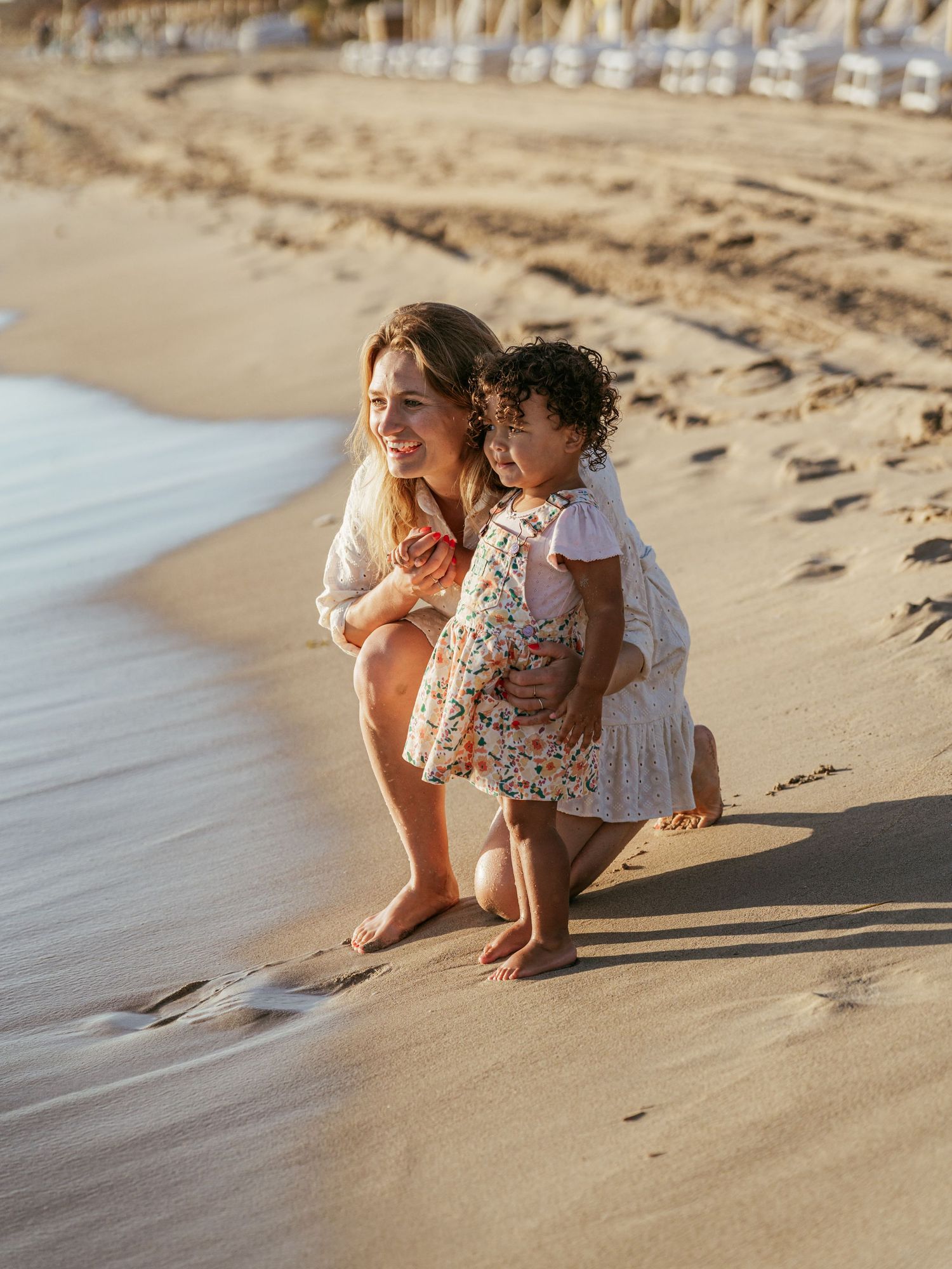 Woman crouching beside a curly-haired toddler on a sandy beach at golden hour.
