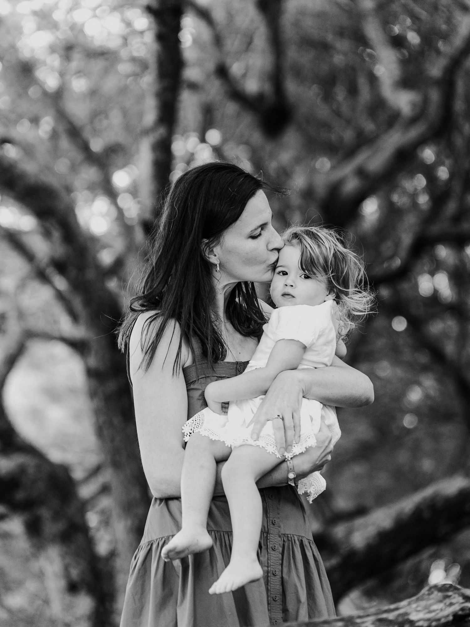 Black and white photo of a mother tenderly kissing her curly-haired toddler held in her arms outdoors.