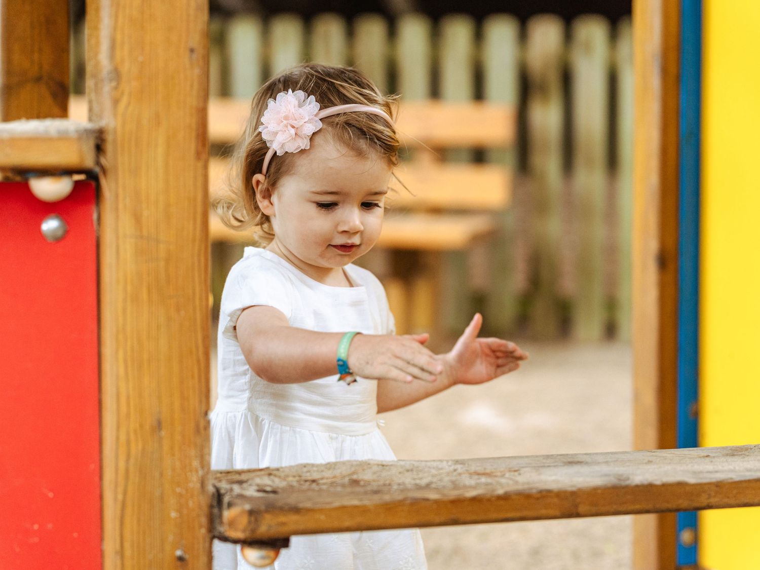 A little girl in a white dress with a pink headband claps hands at a playground.