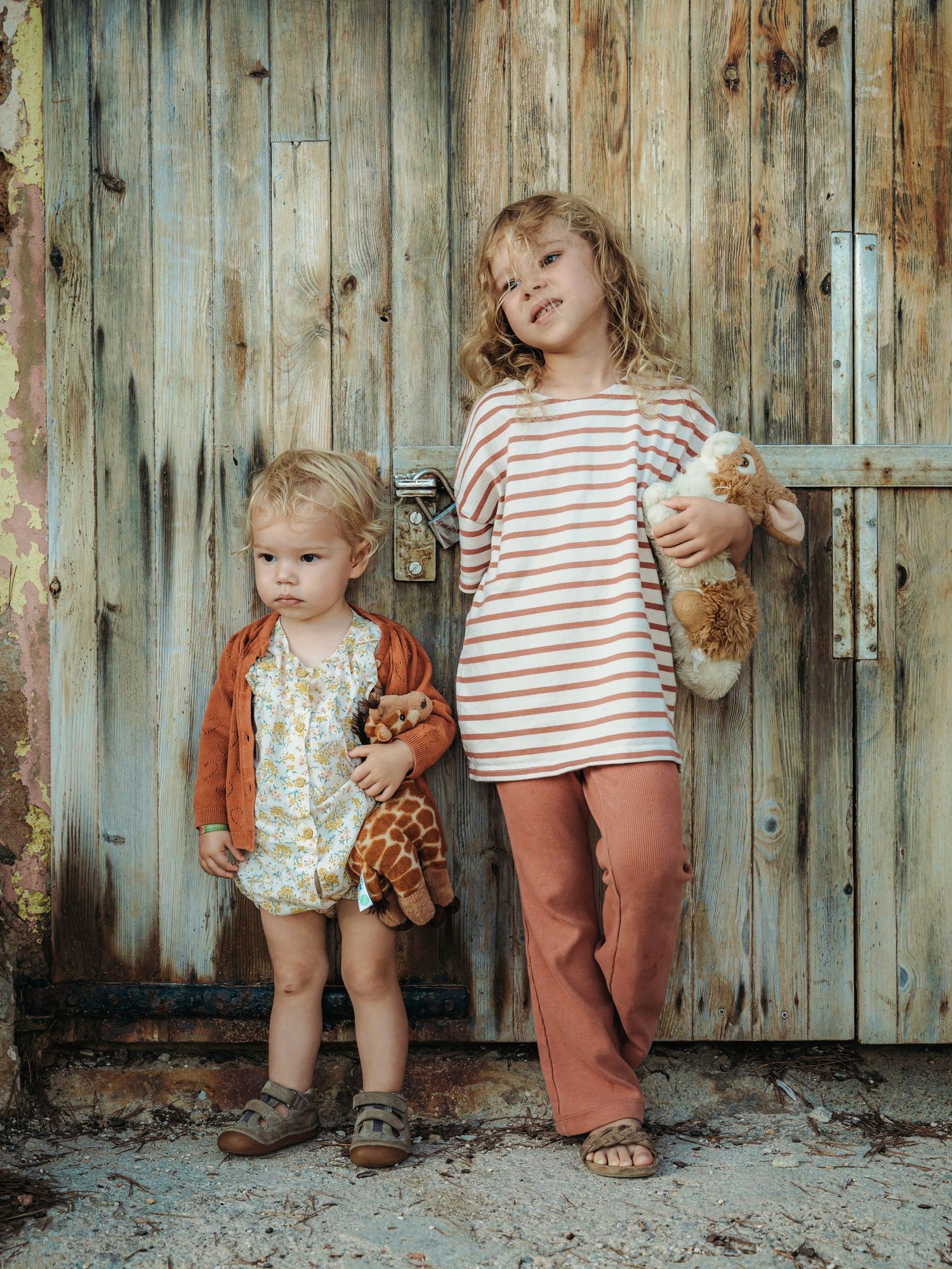 Two young sisters in earth-tone outfits holding stuffed animals beside rustic weathered wooden door.