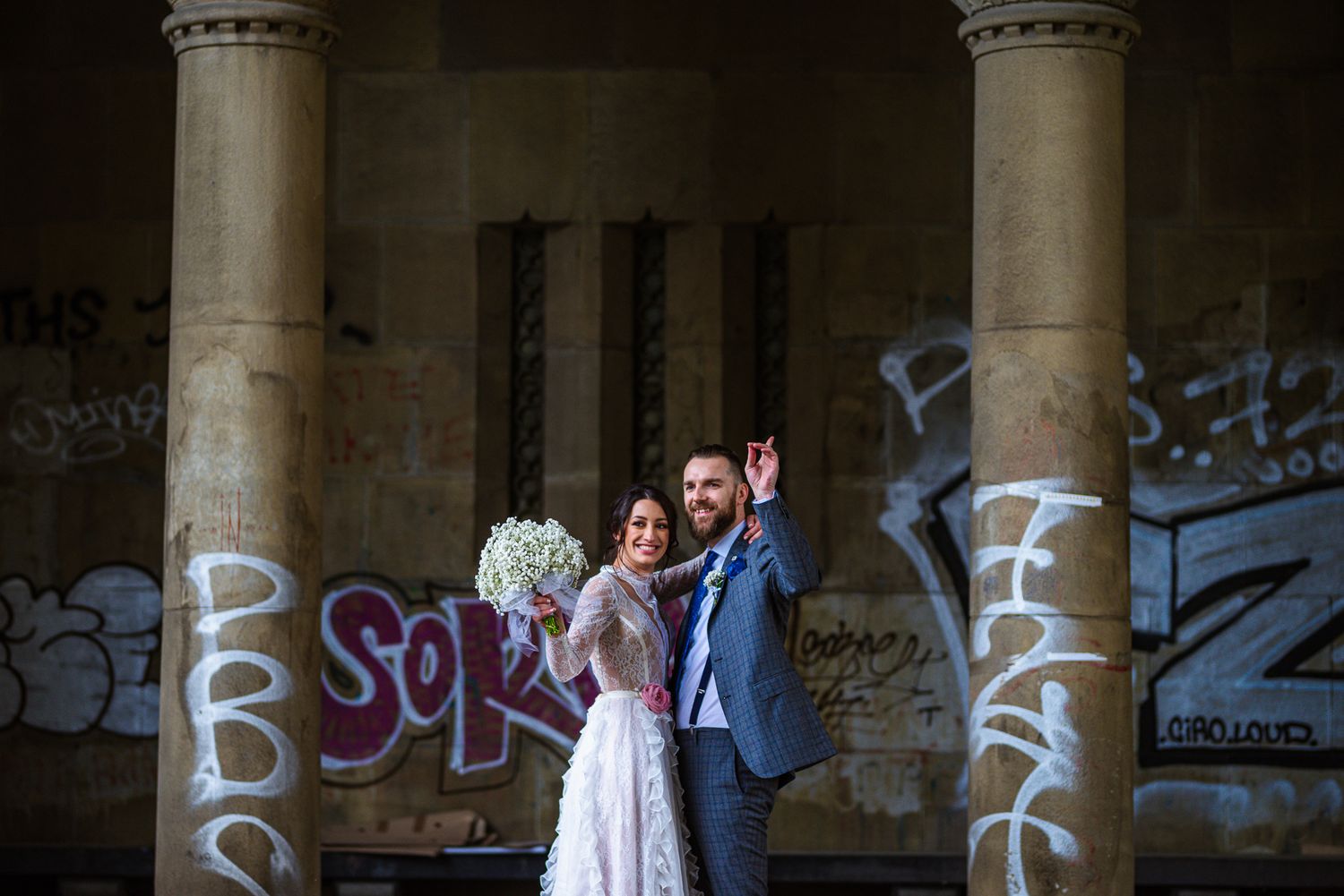 Bride and groom waving joyfully beside graffiti-covered stone columns at an urban wedding location.