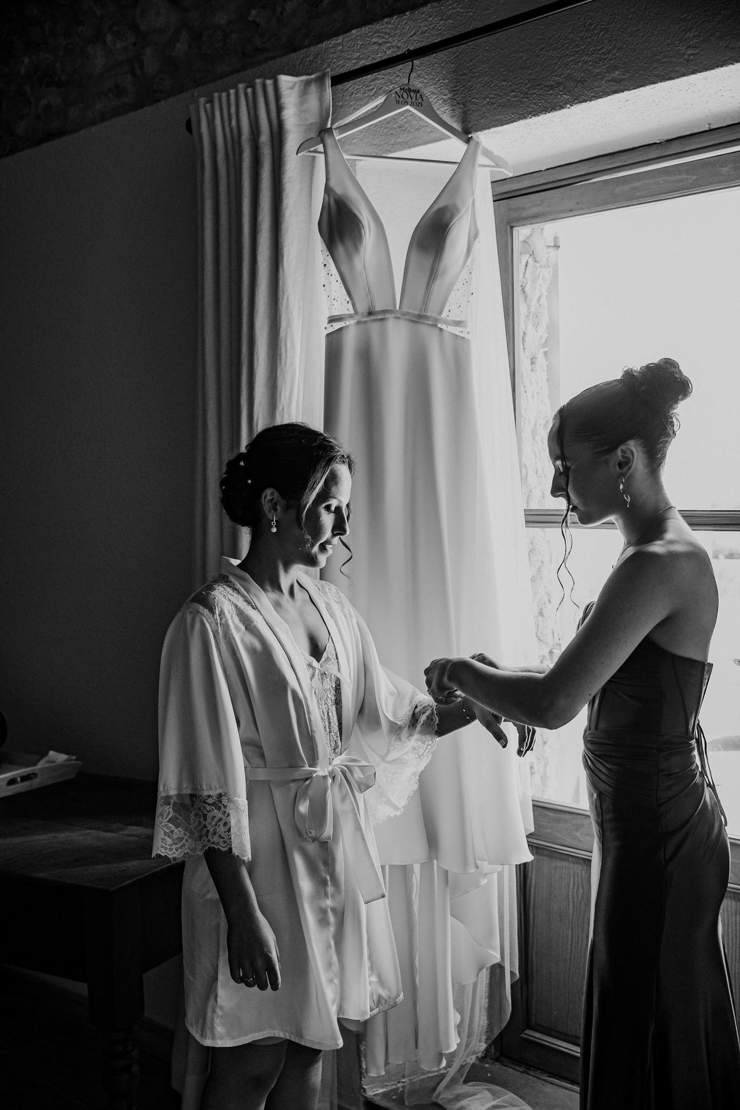 Black and white photo of bridesmaid helping bride prepare near hanging wedding gown.