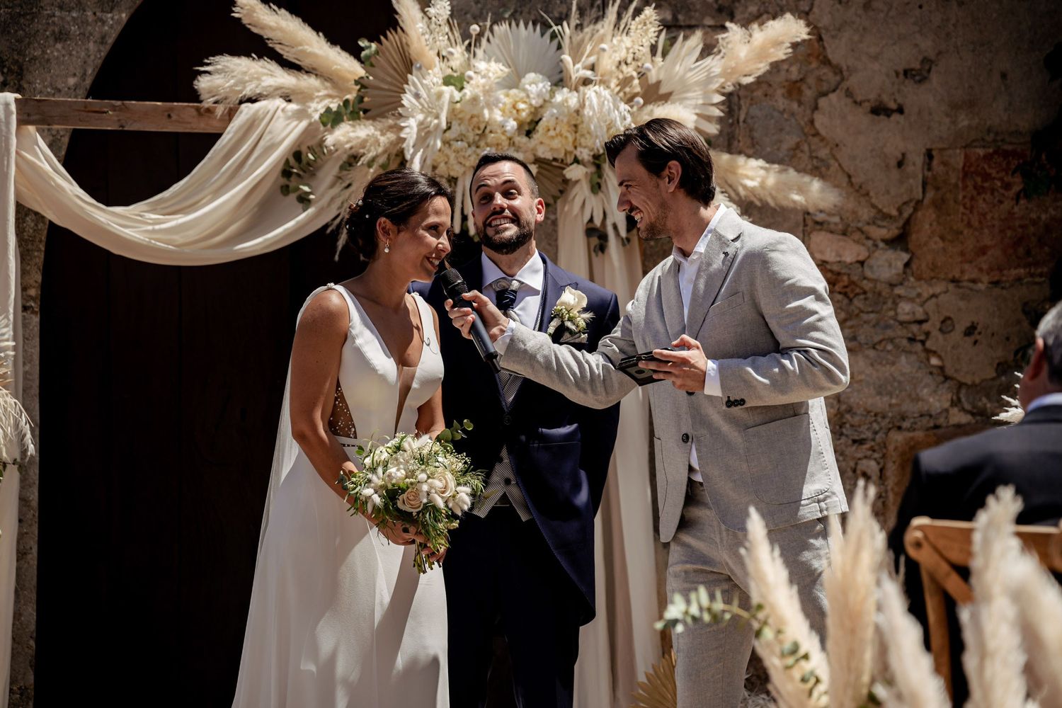Officiant gestures warmly to smiling bride and groom during outdoor boho wedding ceremony.