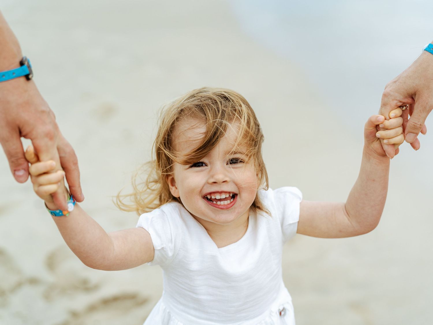 Smiling toddler girl in white outfit held by parents on beach.