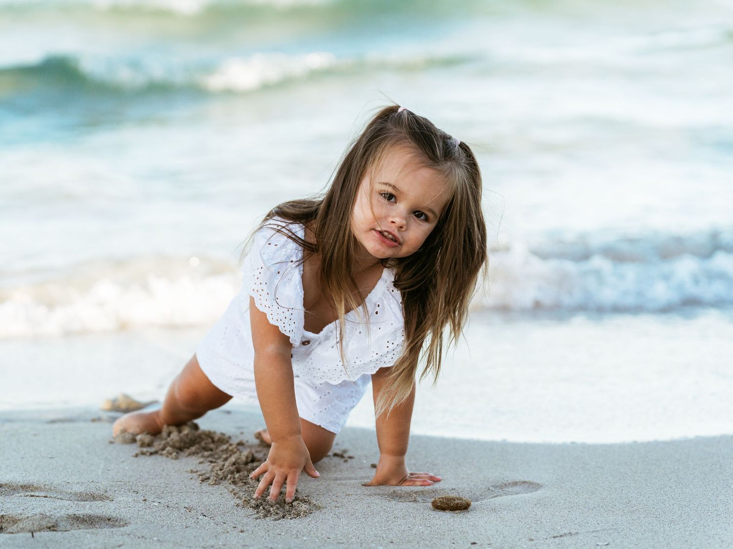 Cute little girl in white outfit crawls on sandy beach with ocean waves behind her.