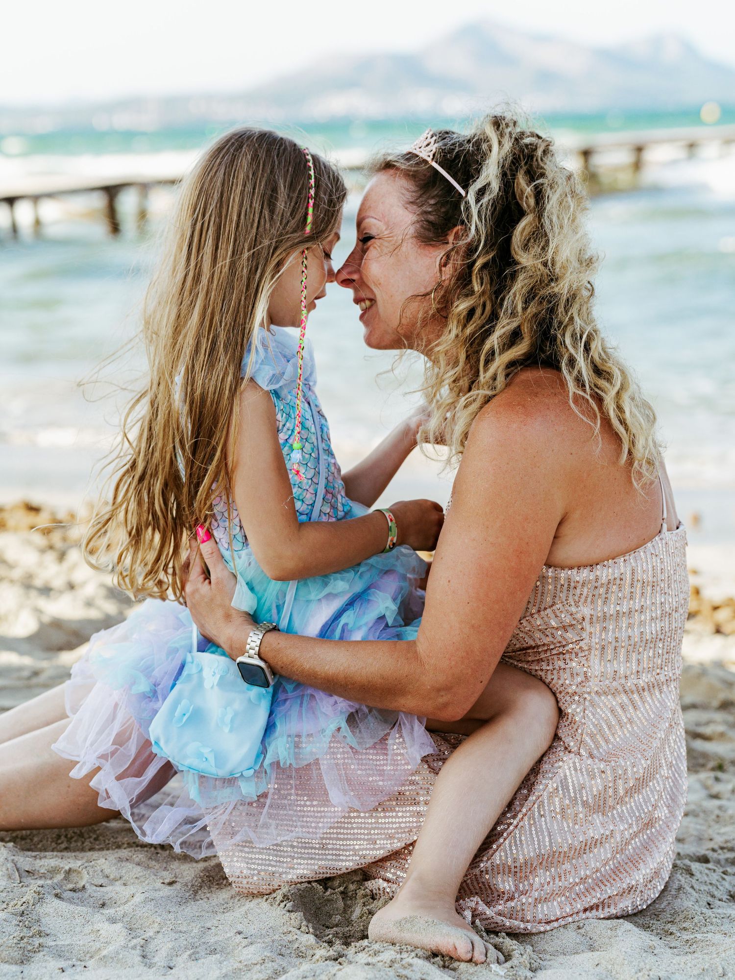 A curly-haired mother and her young daughter touching foreheads affectionately while sitting on a beach.