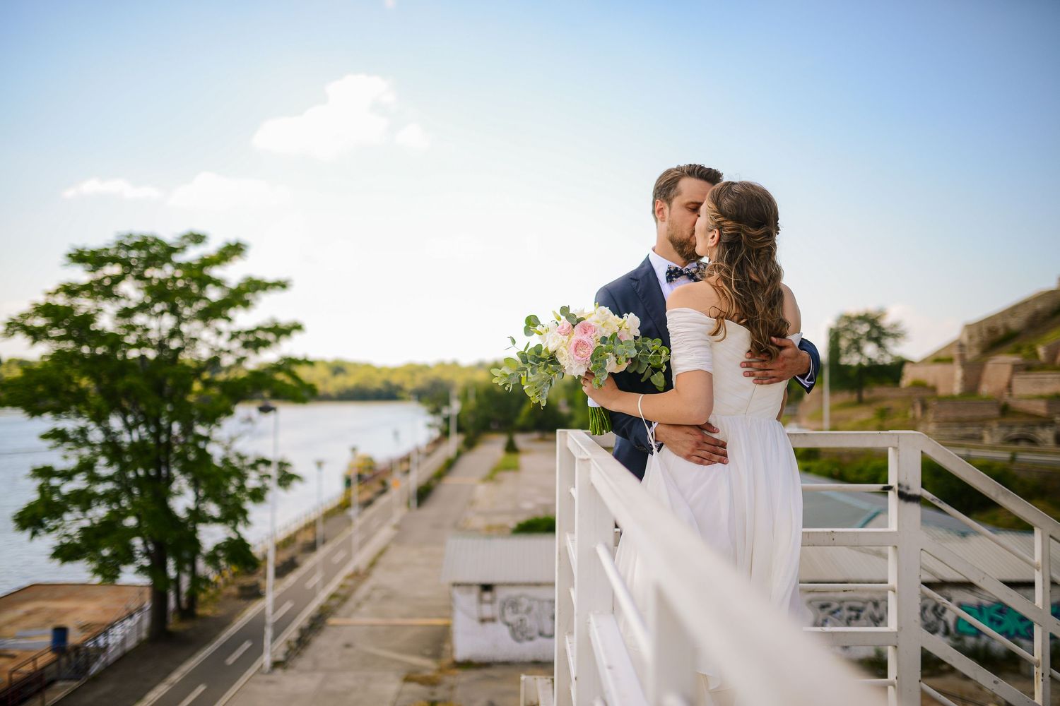 Romantic bride and groom kiss on a waterfront balcony at sunset holding a pastel floral bouquet.