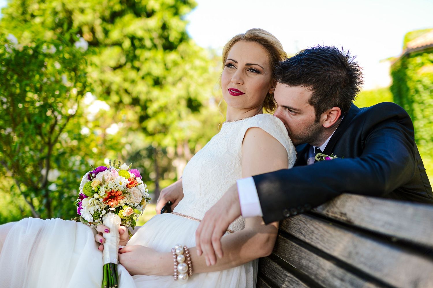 Bride and groom sitting on park bench, groom kissing bride holding colorful bouquet.