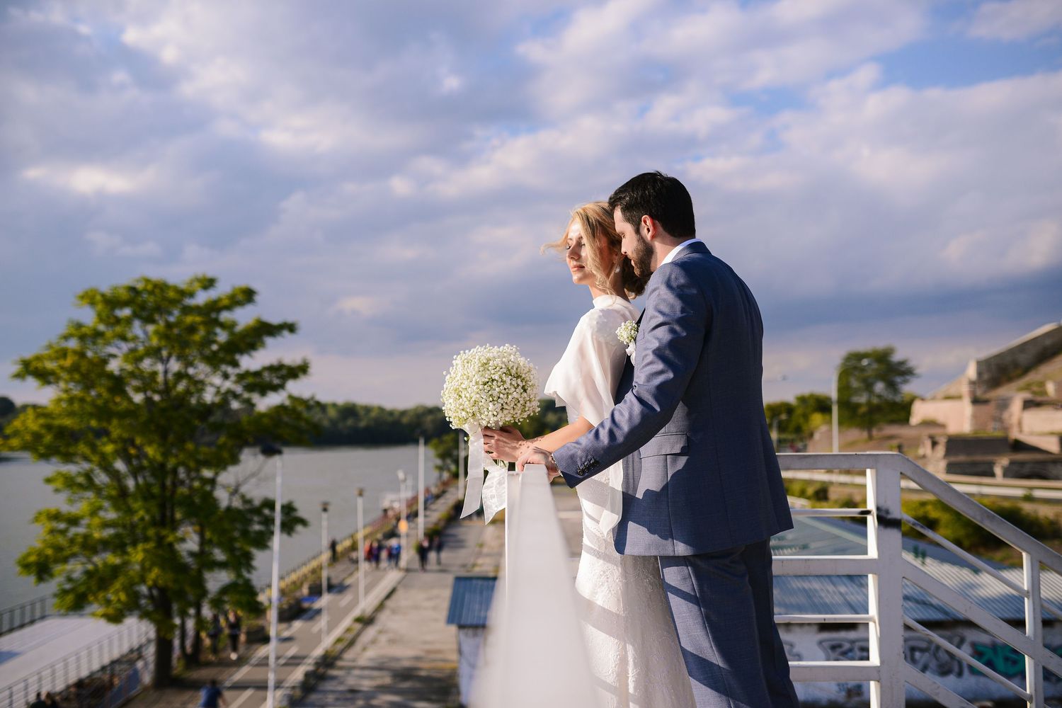 Bride and groom stand together on a waterfront terrace, she holding a white bouquet under a dramatic sky.
