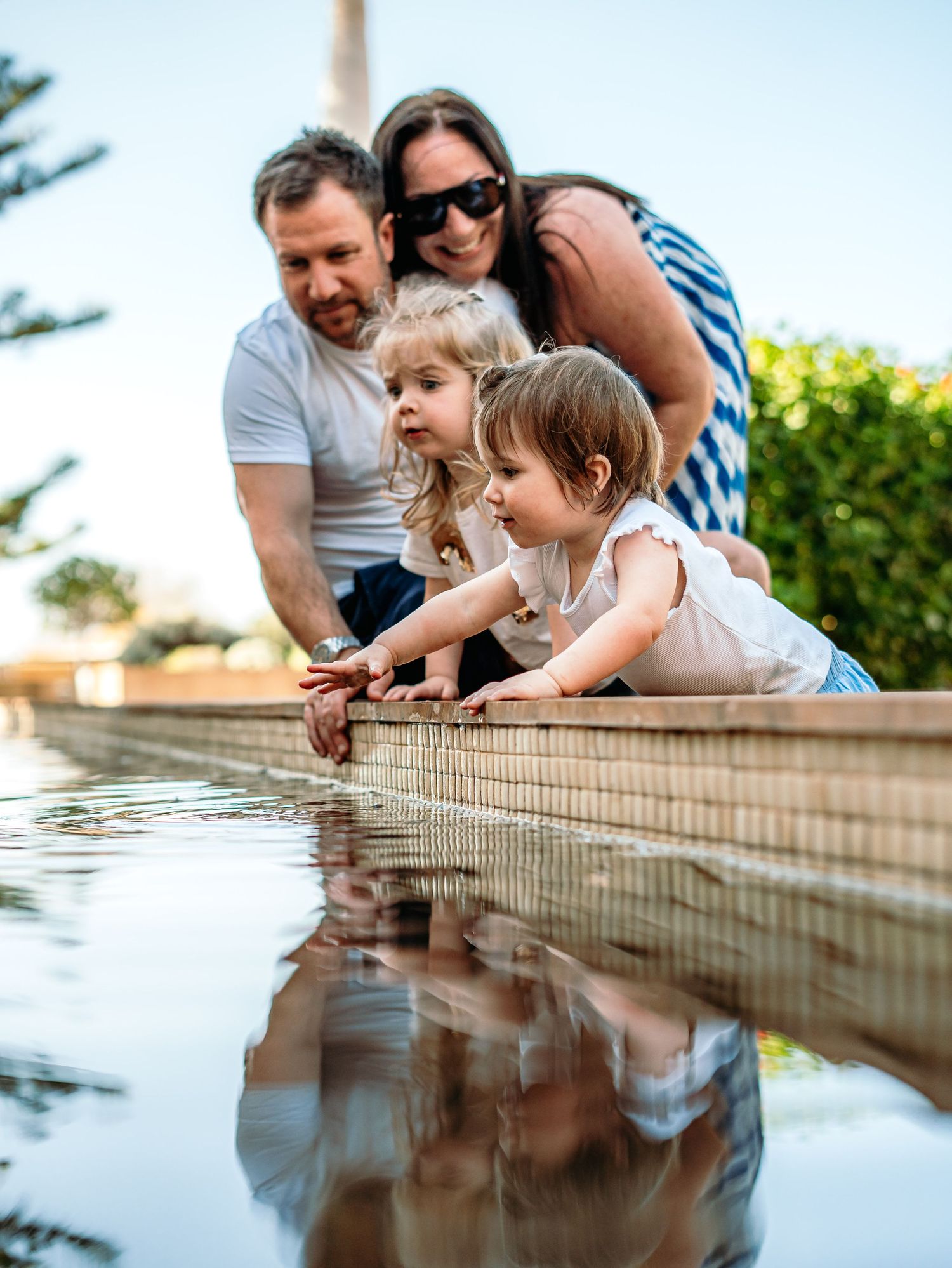 Family of four with two young children touching water in a reflective garden pond on a sunny day.