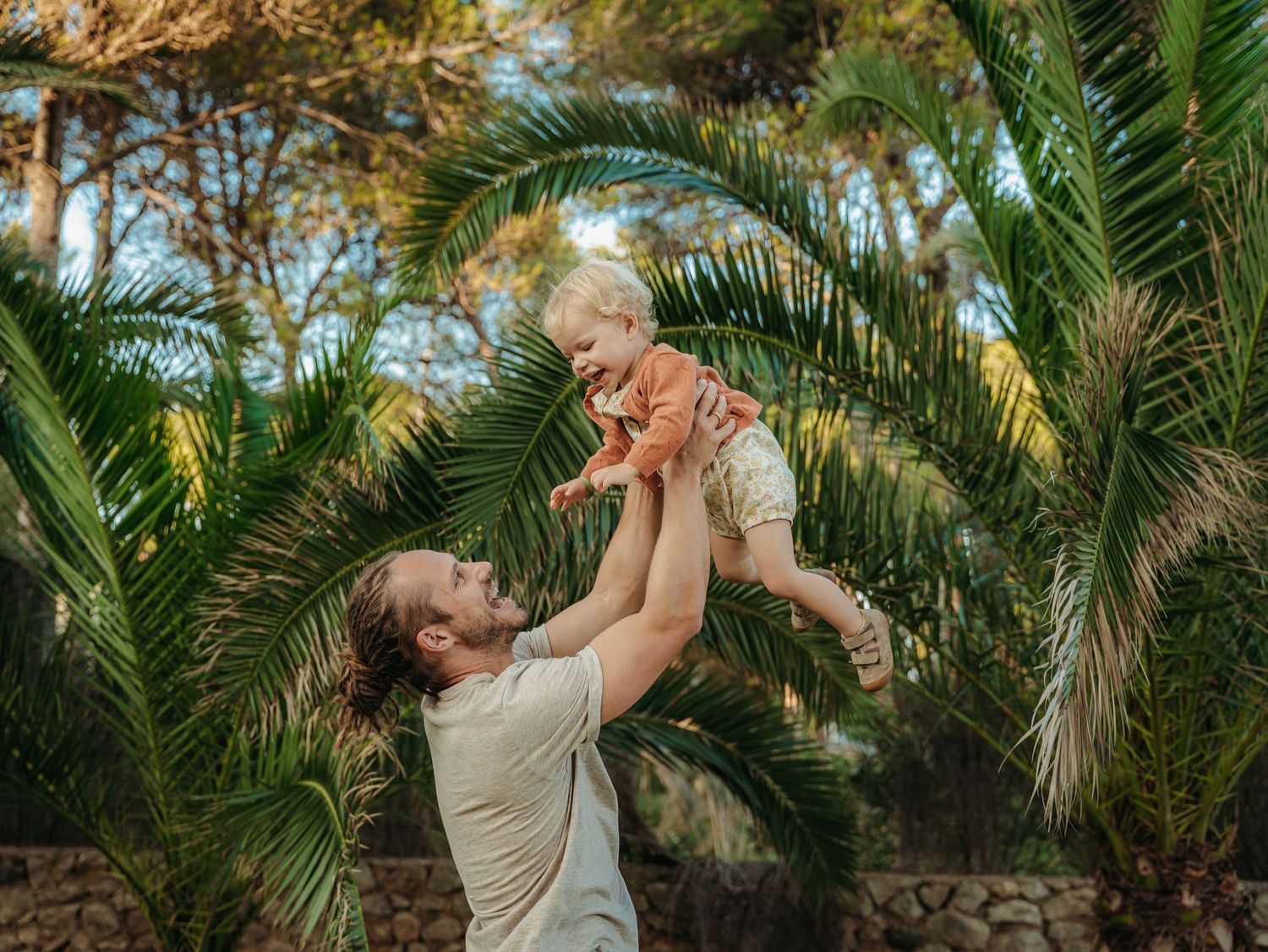 Dad lifting smiling baby high above his head outdoors among lush green palm trees.
