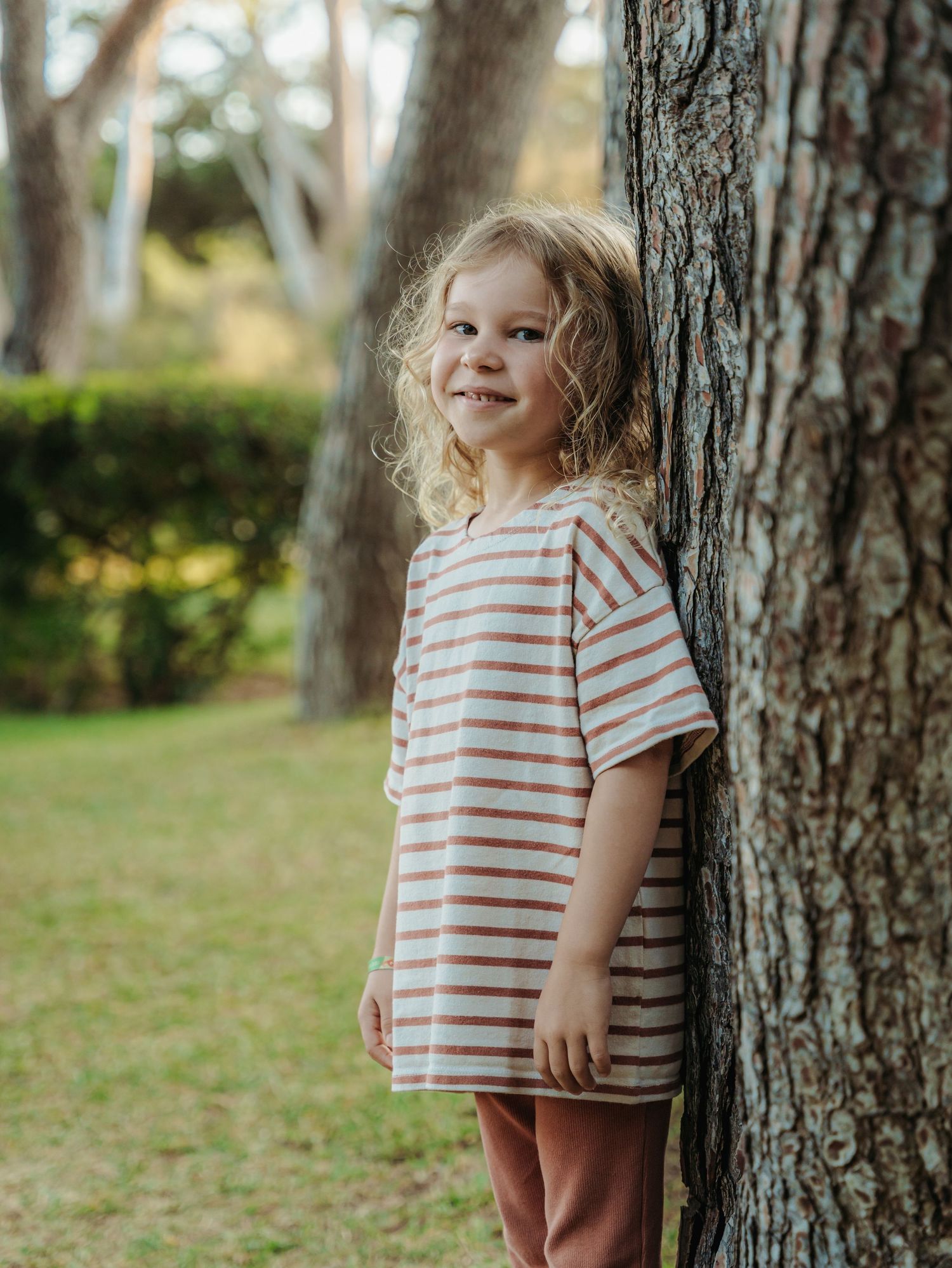 Cute blonde curly-haired girl in striped dress leaning against a tree trunk in a sunny park.