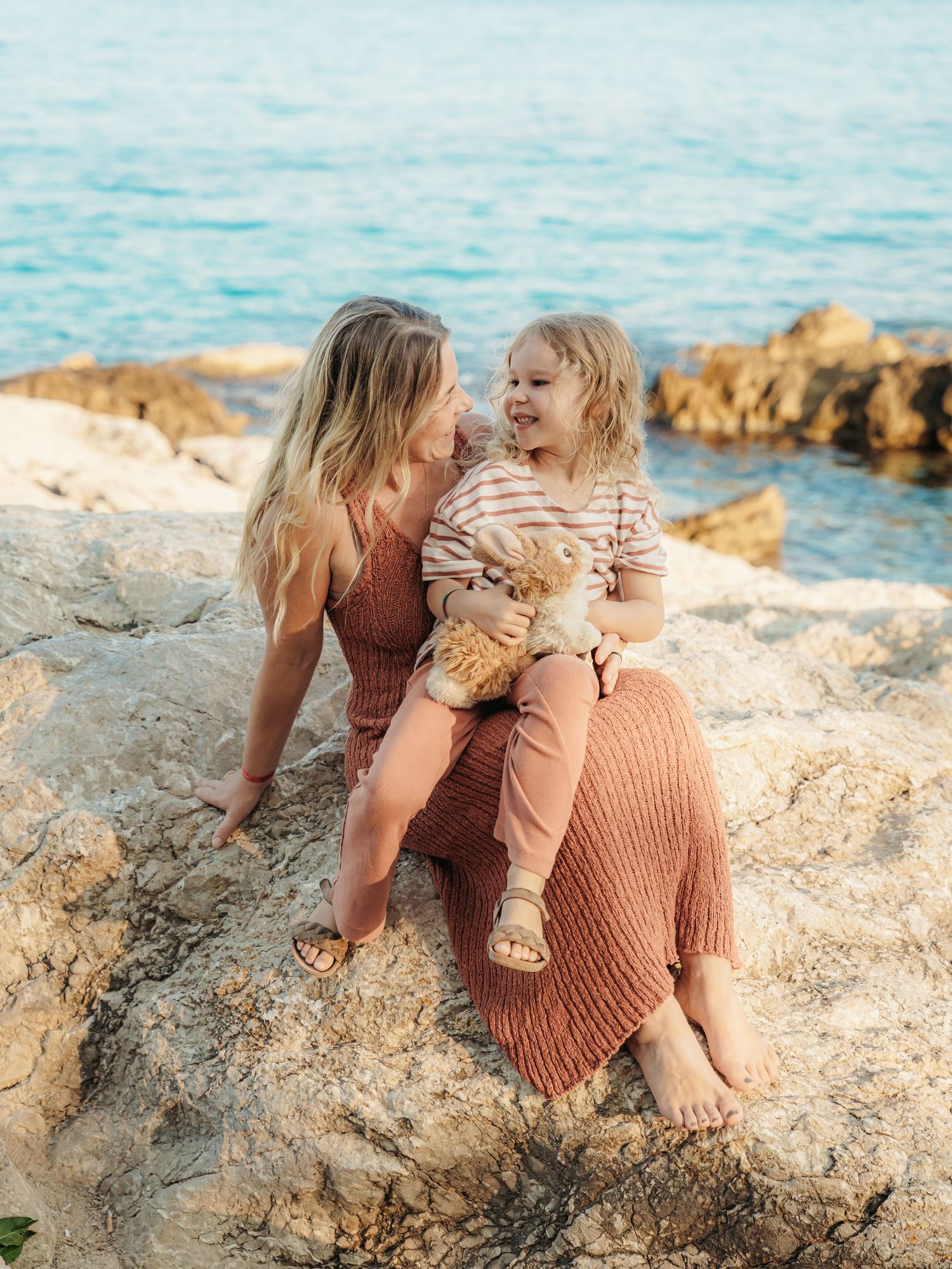 Mother and toddler sitting on coastal rocks by turquoise sea, child holding stuffed animal.
