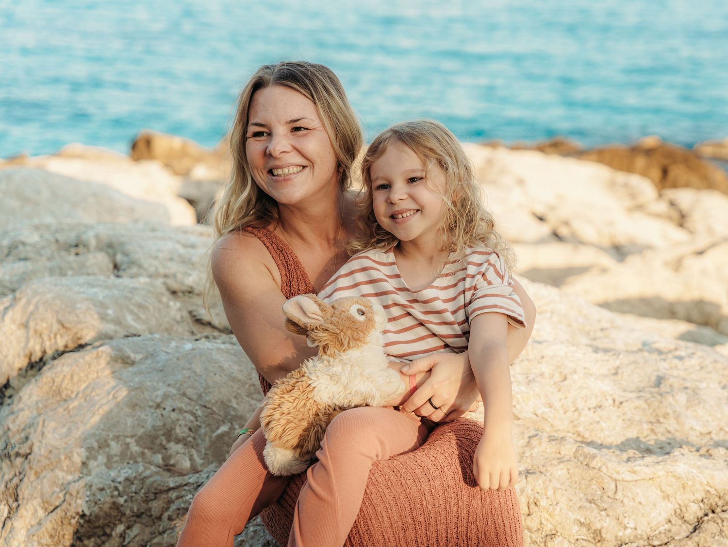 Smiling mother and young daughter posing together on rocky beach with ocean behind.
