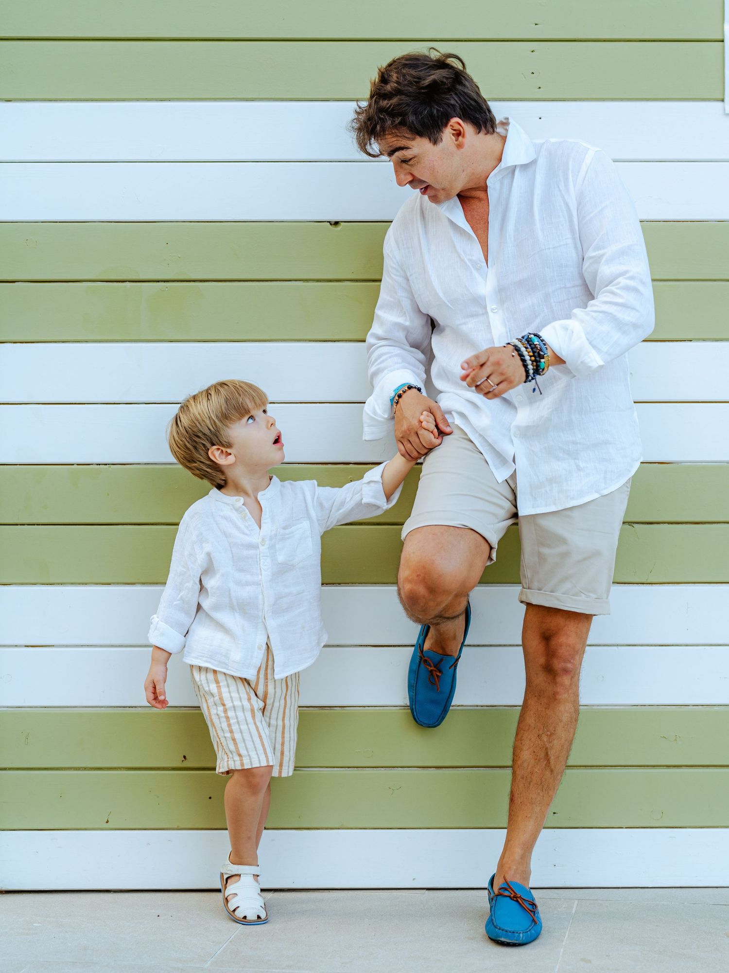 A father in white linen leans down to hold hands with his young son against a wall.