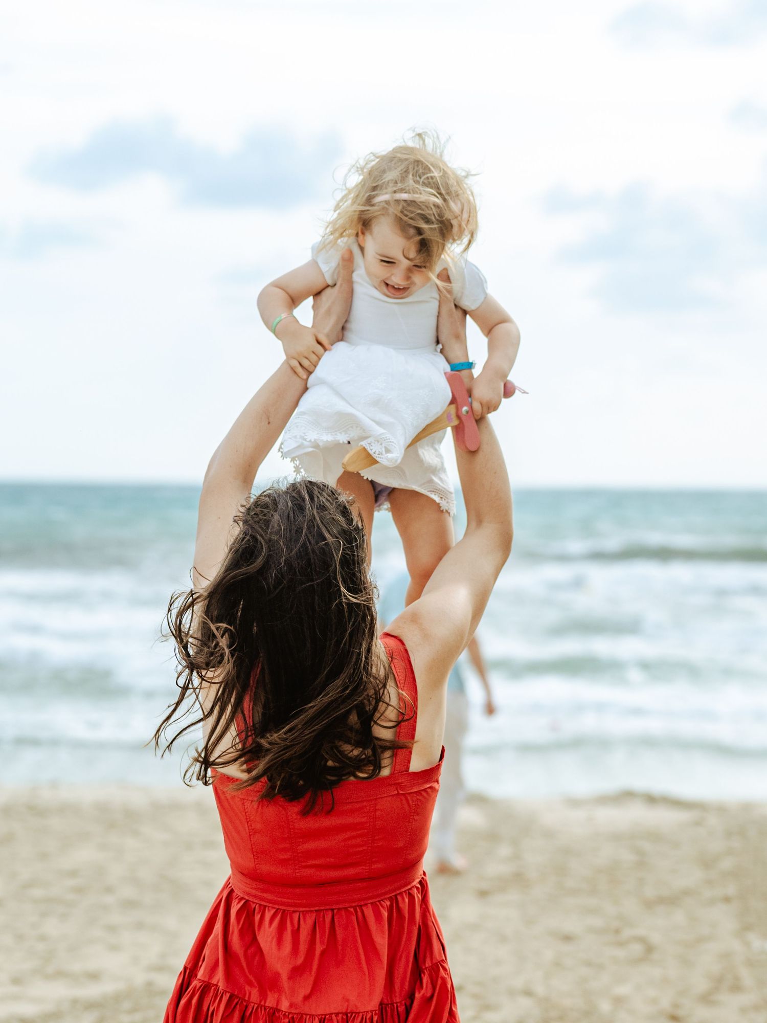 Mother in red dress lifting laughing toddler high in the air on a sunny beach.