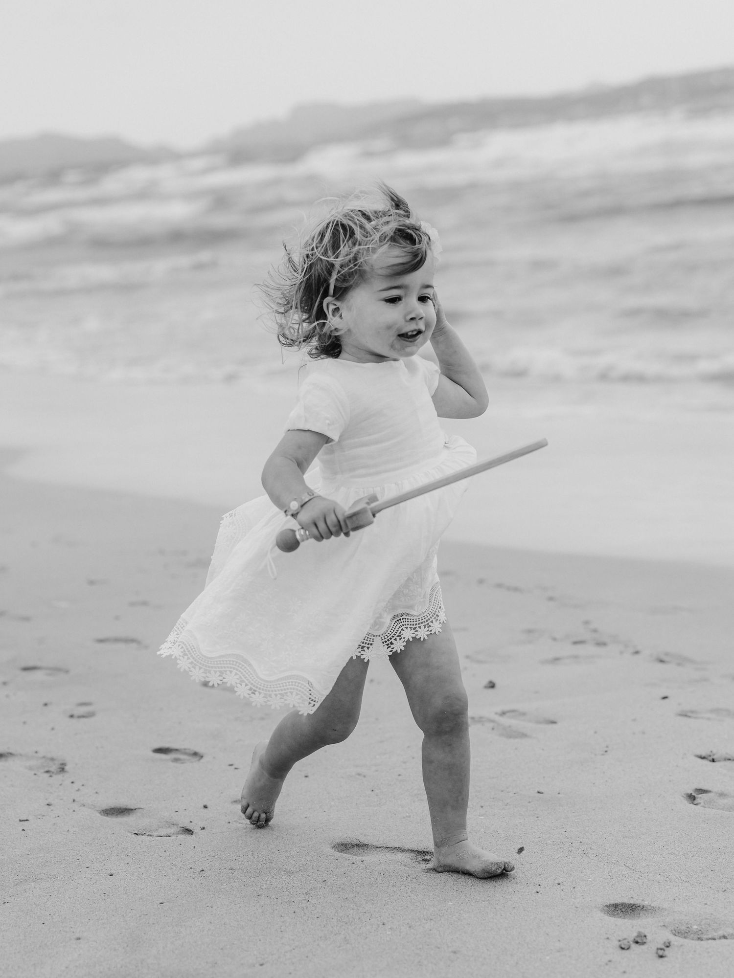 Black and white photo of toddler girl in white dress playing on beach.