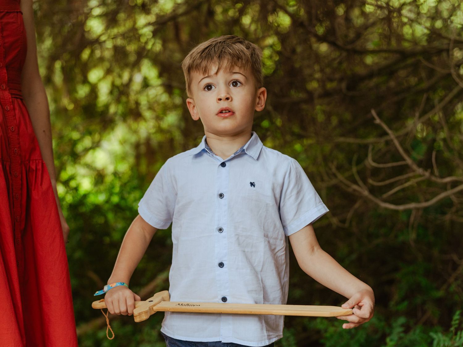 Young boy in light blue shirt holding wooden sword standing outdoors with green trees behind.