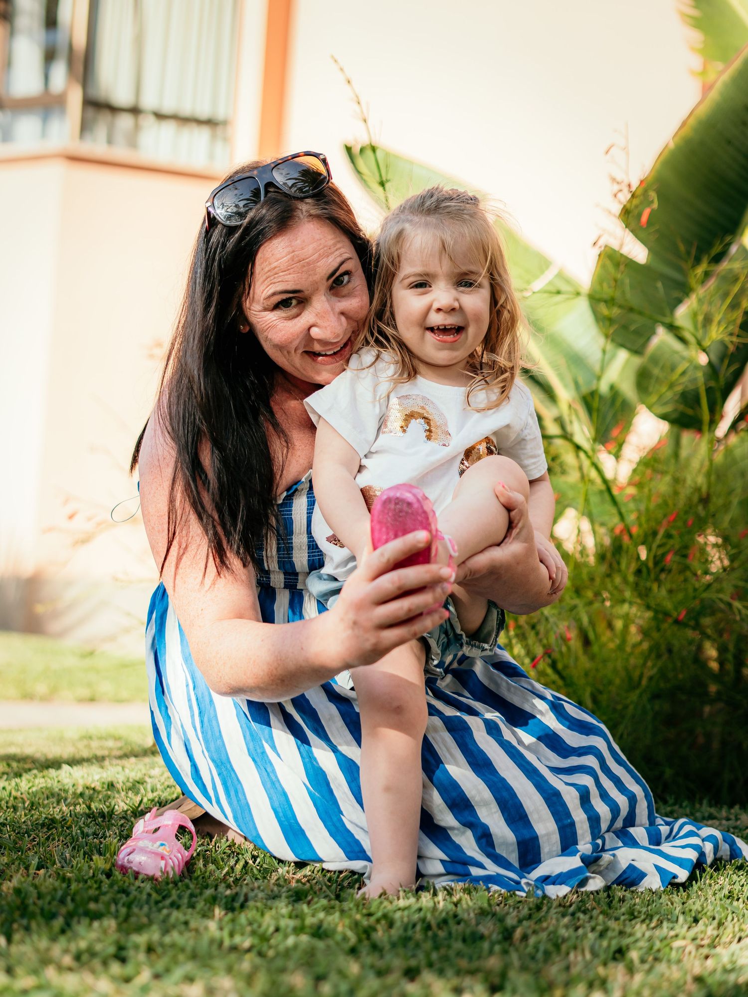 Happy mother in striped dress sitting on grass holding her smiling young daughter outdoors.
