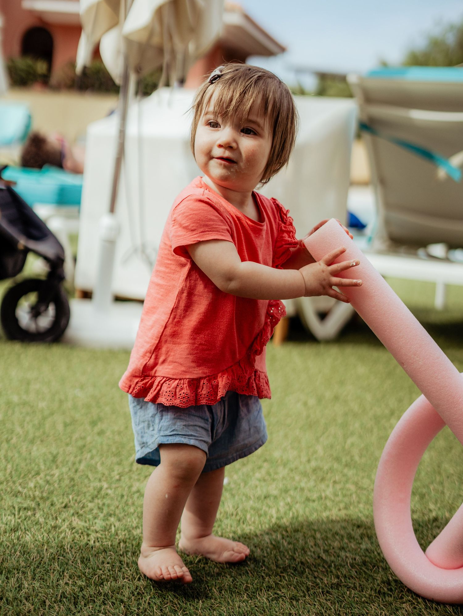 Toddler girl in red top and denim shorts holding a pink flamingo toy on green grass.