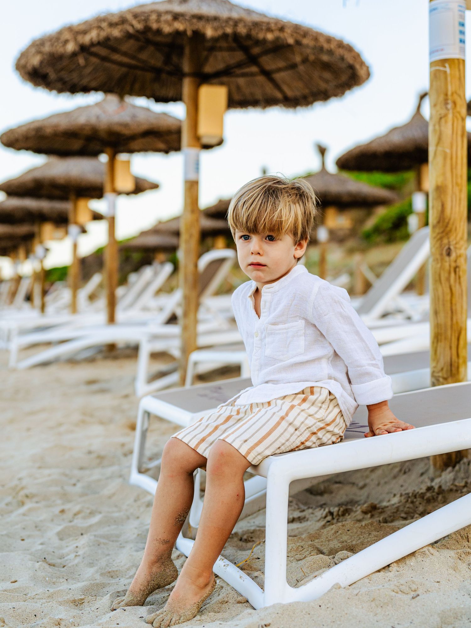 Young boy in white shirt and striped shorts sitting on beach lounger under straw umbrellas.