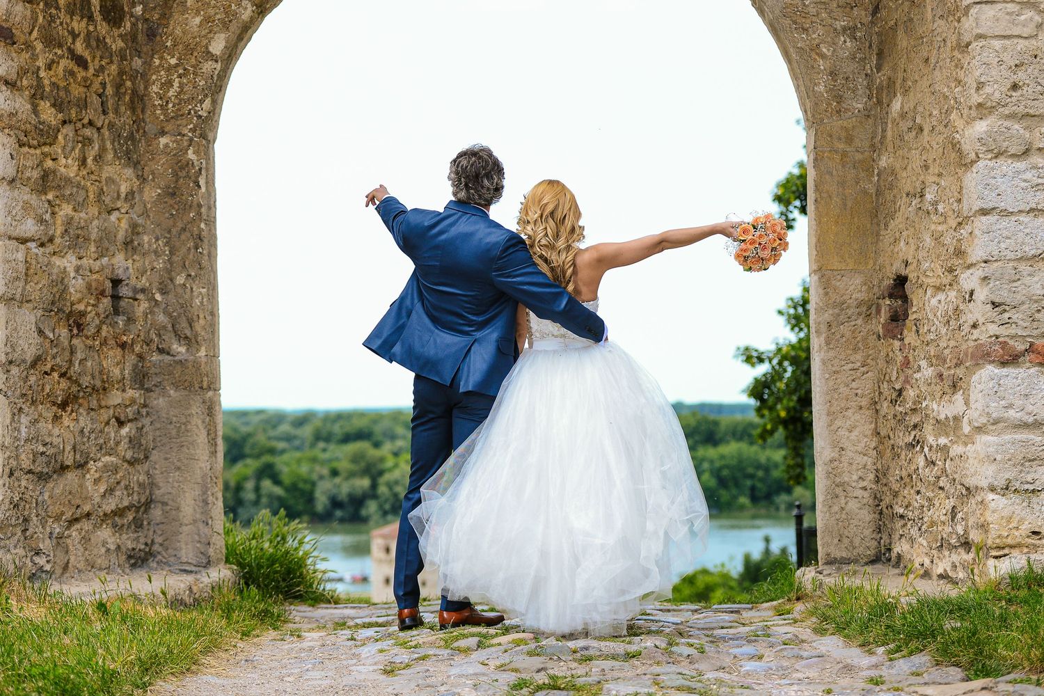 Bride and groom stand in a stone archway overlooking a scenic river, arms outstretched in a joyful wedding pose.