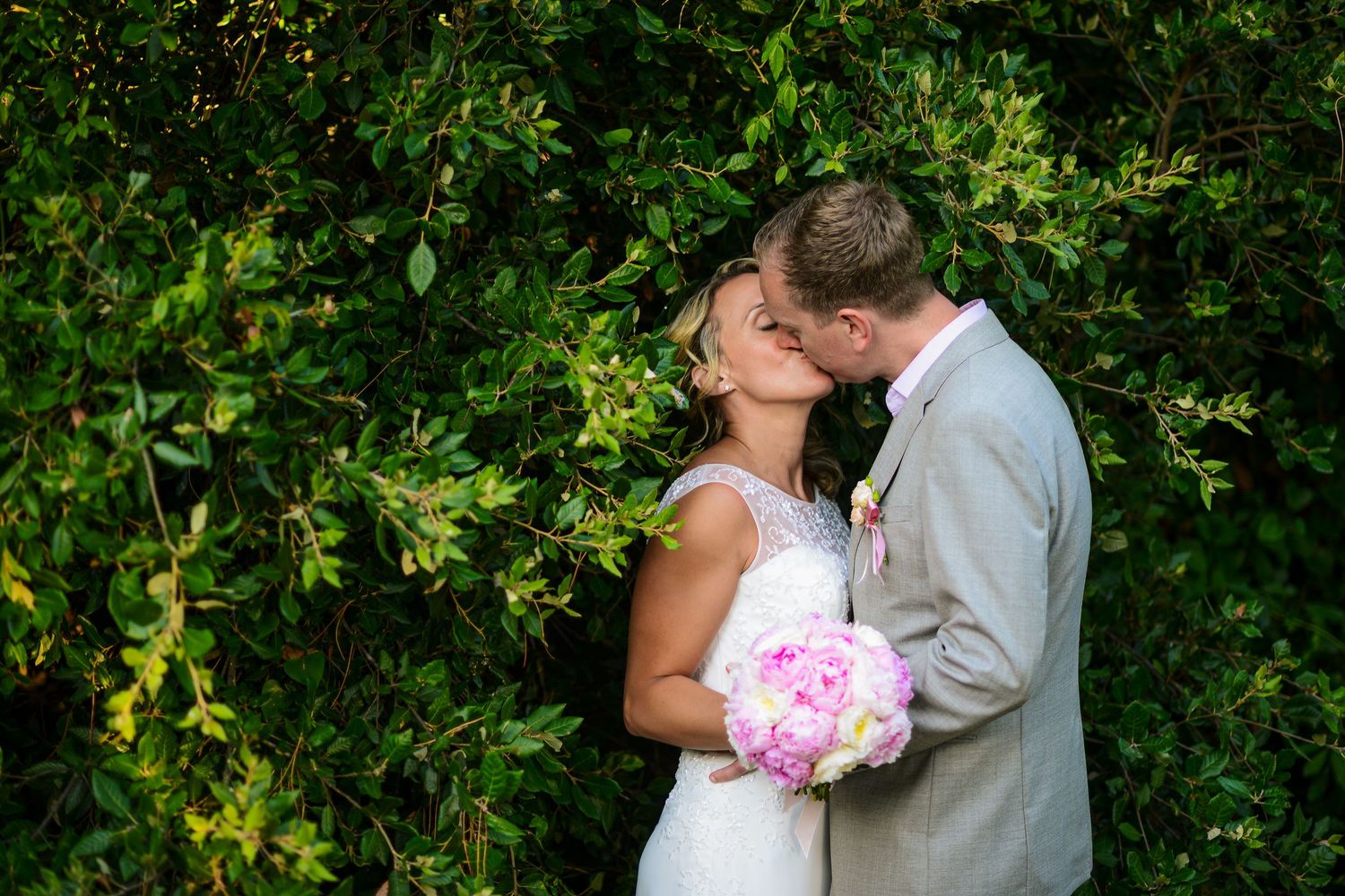 Bride and groom sharing a kiss in front of a lush green hedge wall.