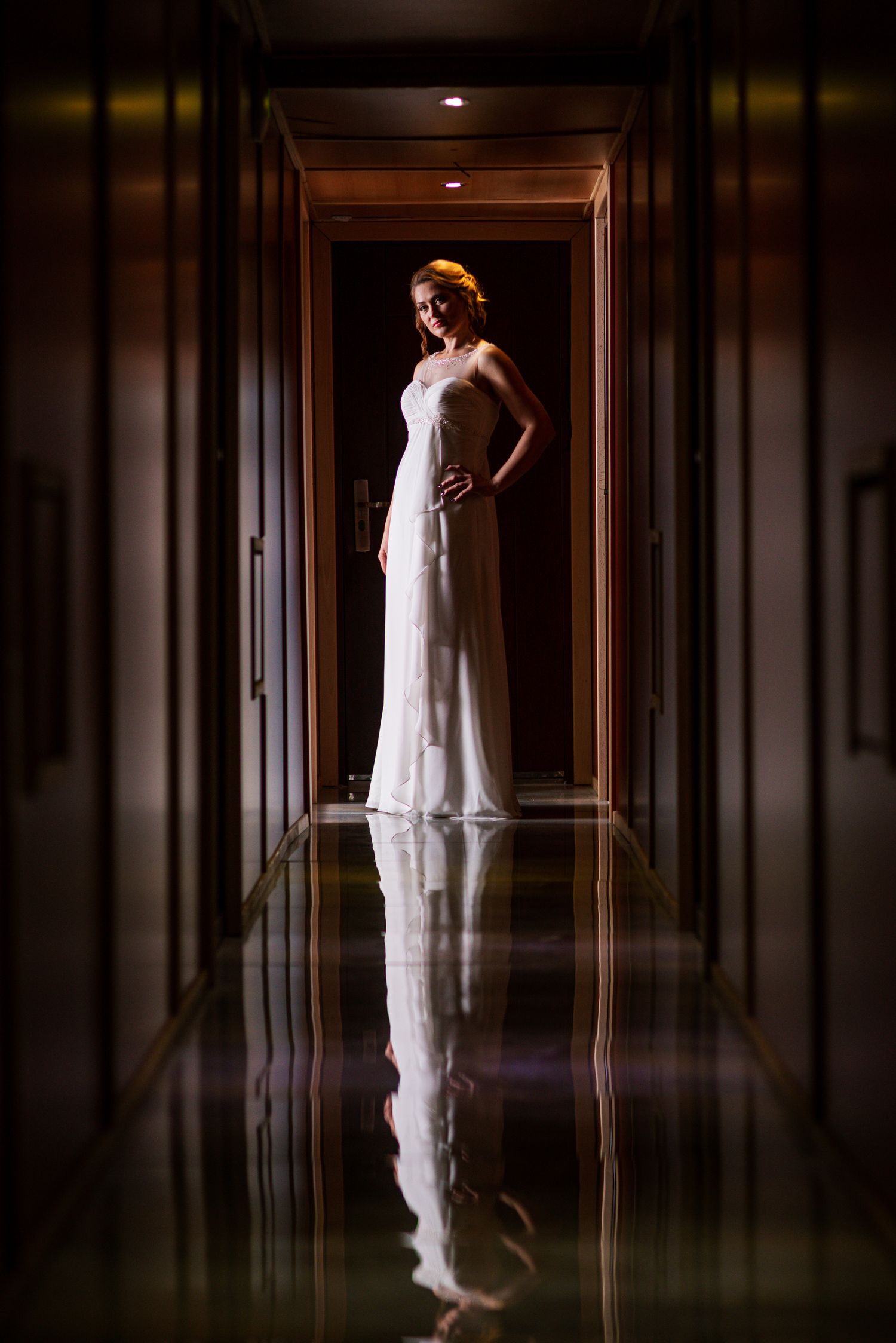 Elegant bride in white gown standing in a dramatically lit hotel corridor with floor reflection.
