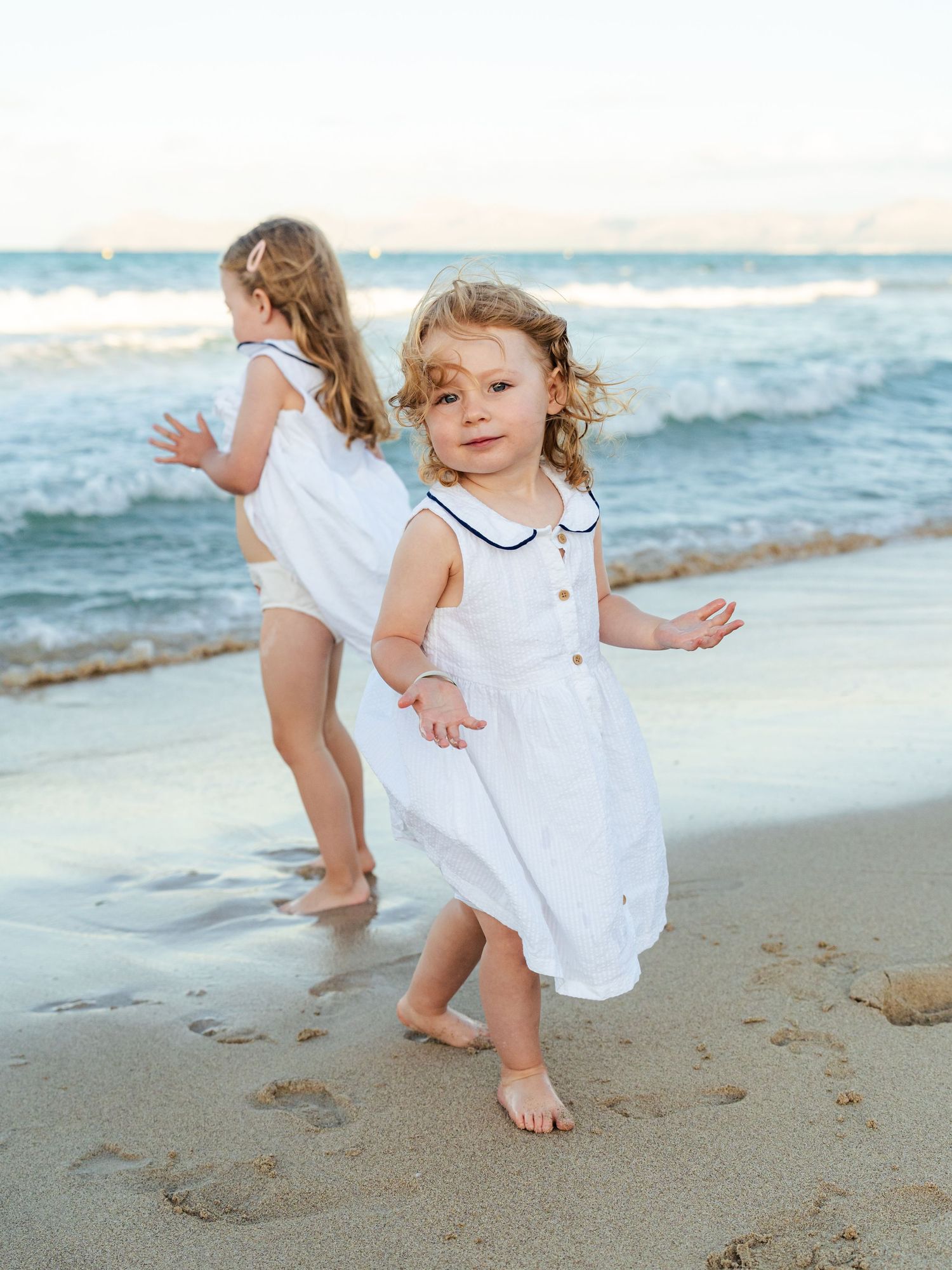 Curly-haired toddler girl in white dress playing on beach near ocean waves.