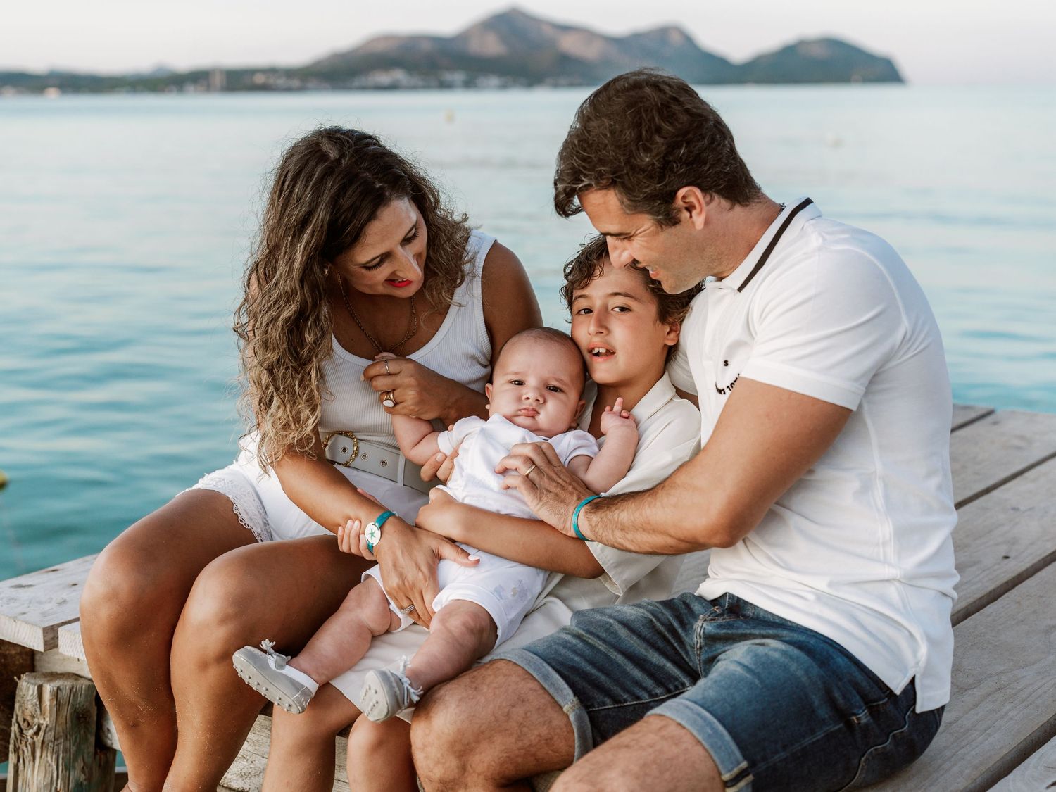 Happy family of four sitting together on a wooden dock enjoying a scenic waterfront view.