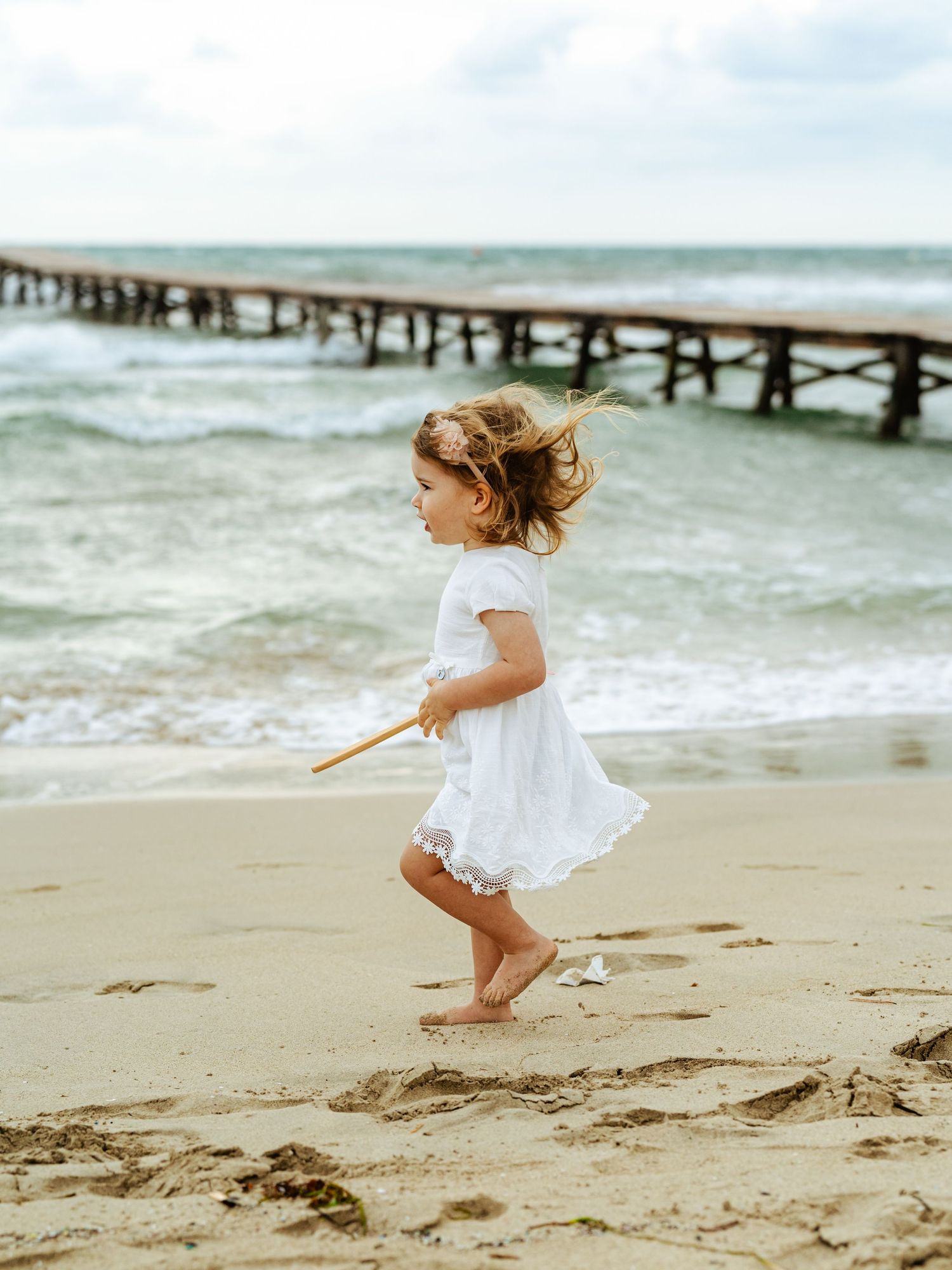 Little girl in white lace dress running barefoot on sandy beach with ocean waves and pier behind.