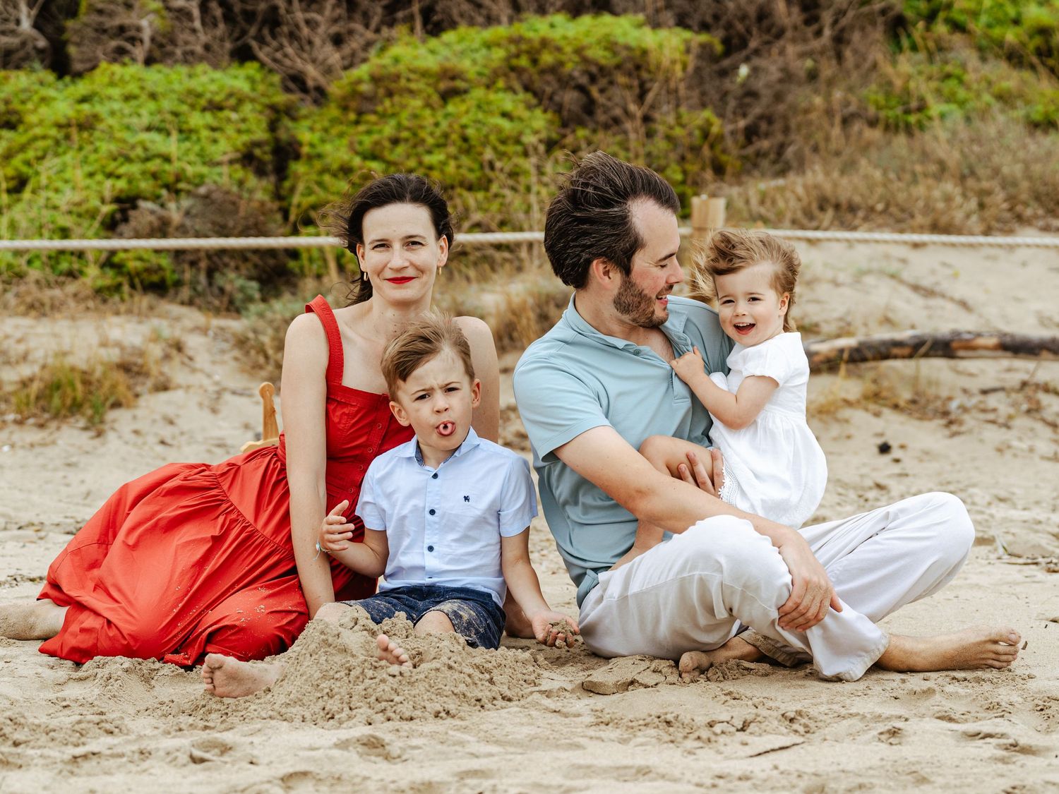 Loving family of four laughing and sitting together on a sandy beach outdoors.