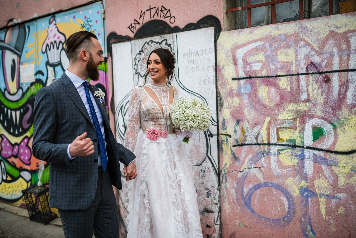 Bride in lace gown and groom walking hand in hand past colorful graffiti wall on wedding day.