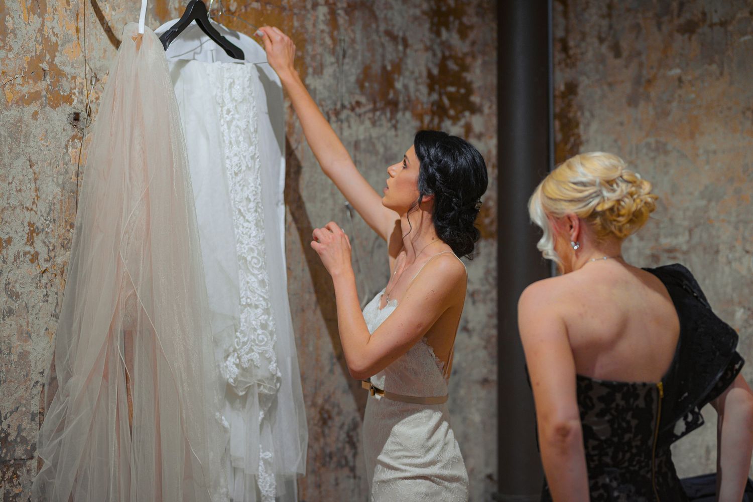 Bride and blonde bridesmaid examine a lace wedding dress hanging on a wall.