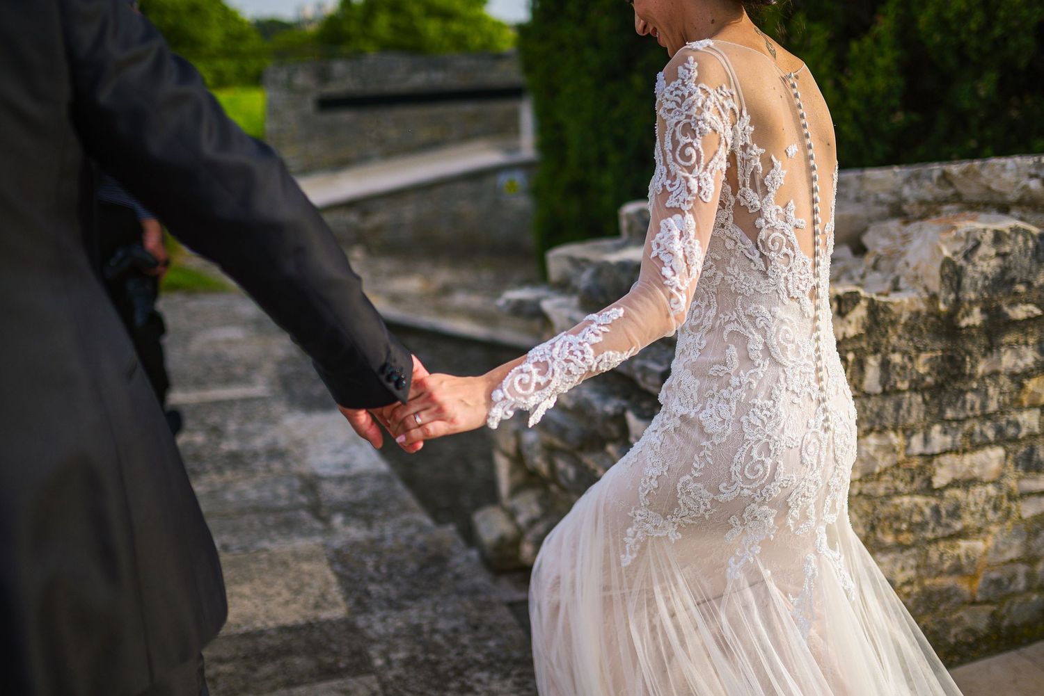 Close-up of bride in intricate lace gown holding groom's hand on stone steps outdoors.