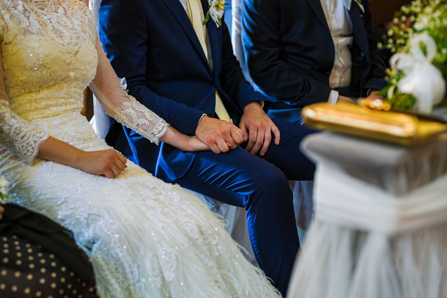 Bride and groom in blue suit holding hands during wedding ceremony while seated together.