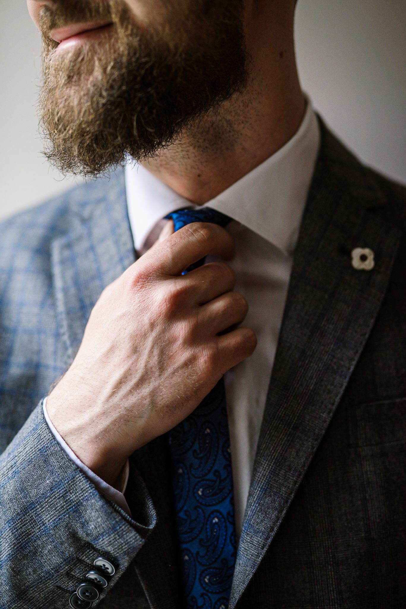 Bearded groom in gray suit adjusting his blue paisley tie before the wedding ceremony.