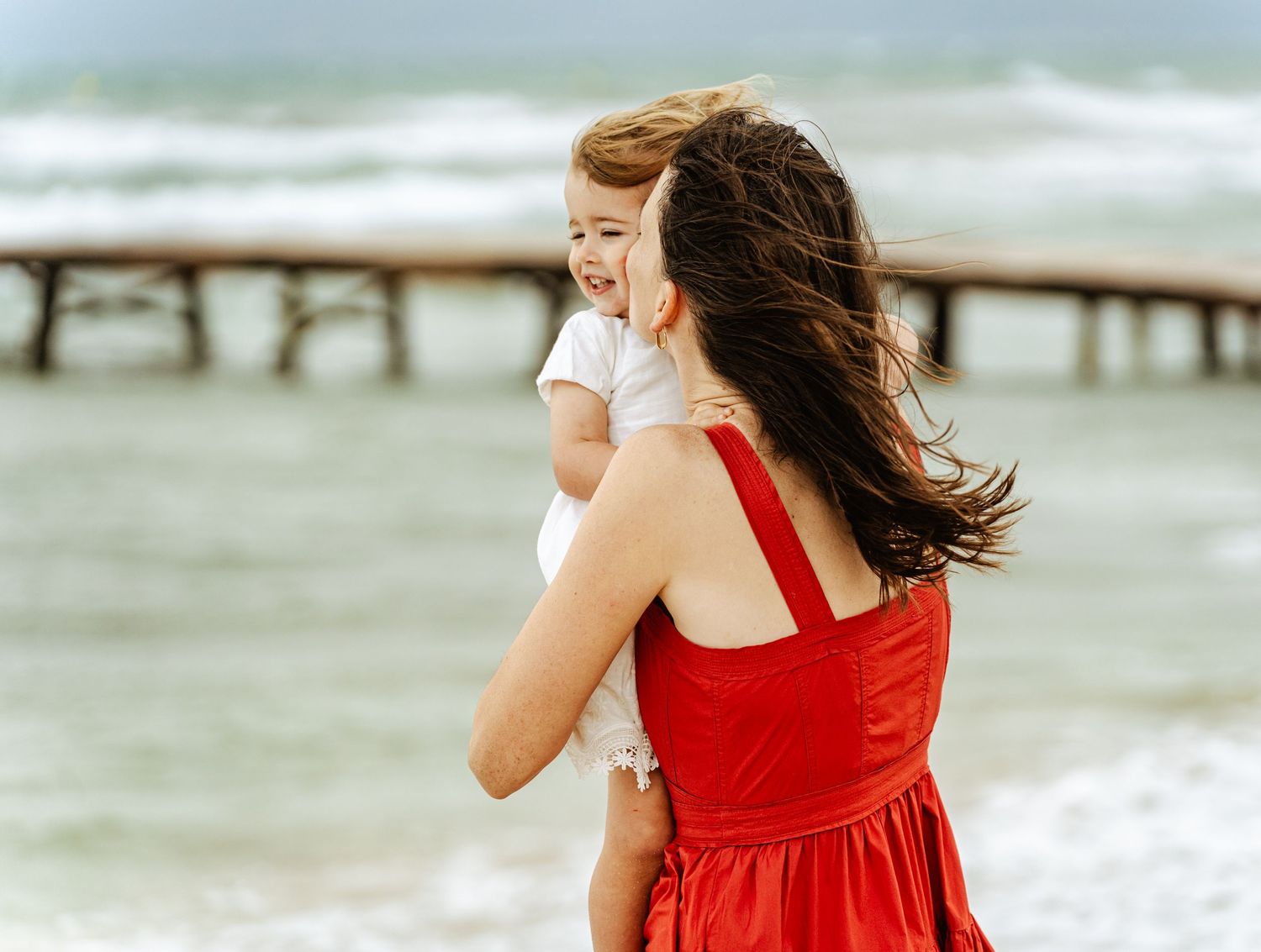 Mother in red dress holding smiling toddler on breezy beach with pier.