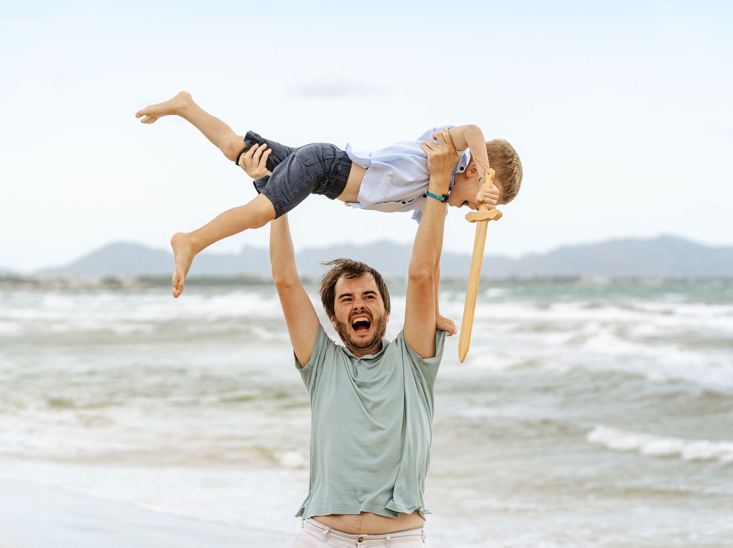 Excited father raising child overhead on beach with ocean waves behind them.