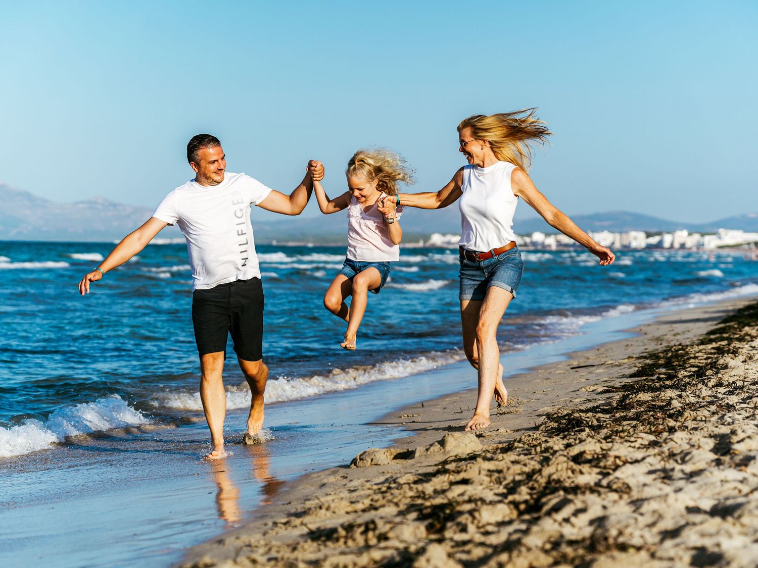 Family of three joyfully running and playing on a sunny beach, with parents swinging their child between them.