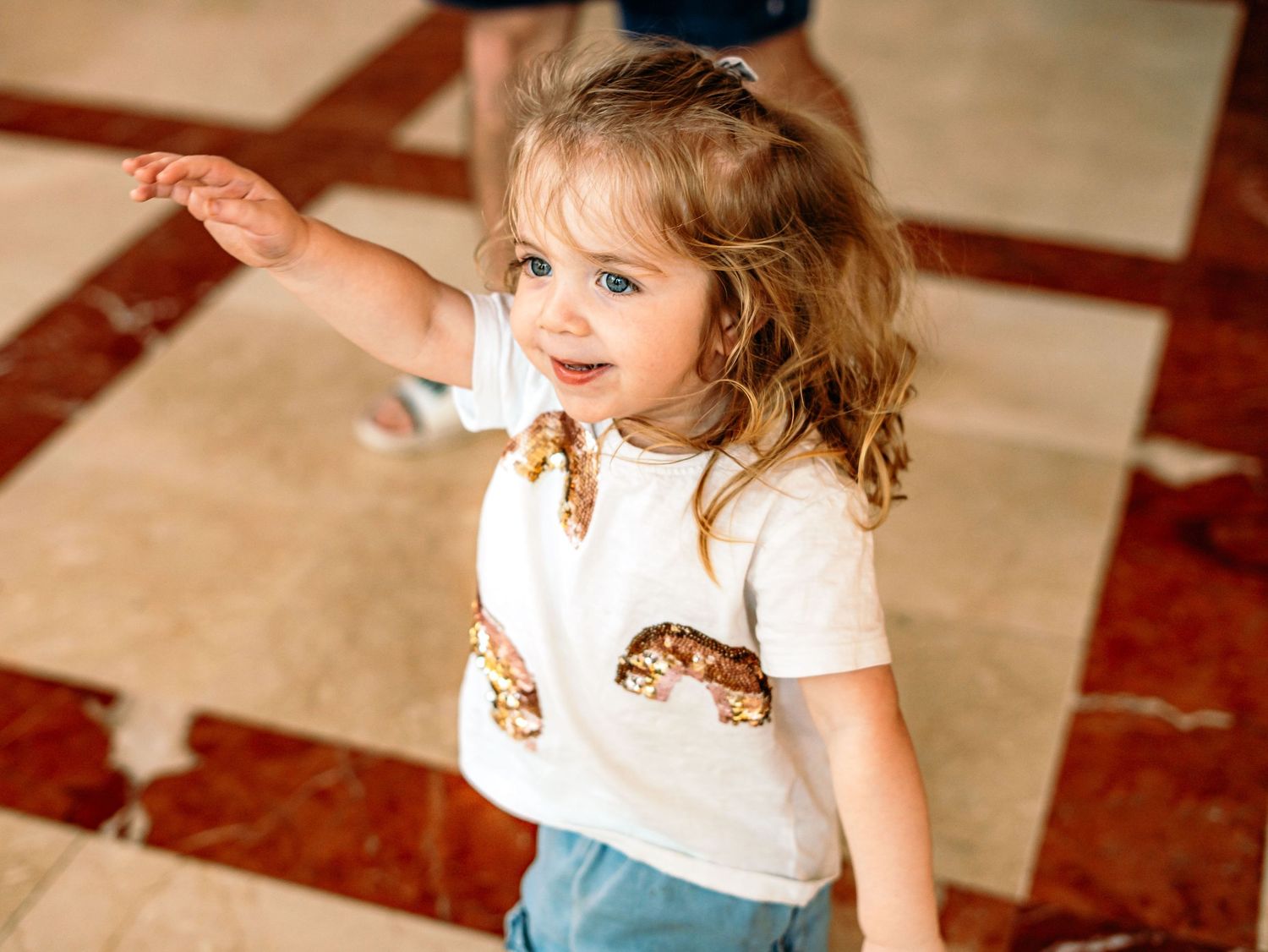 Adorable toddler girl in sequin t-shirt raising arm while standing on marble floor indoors.